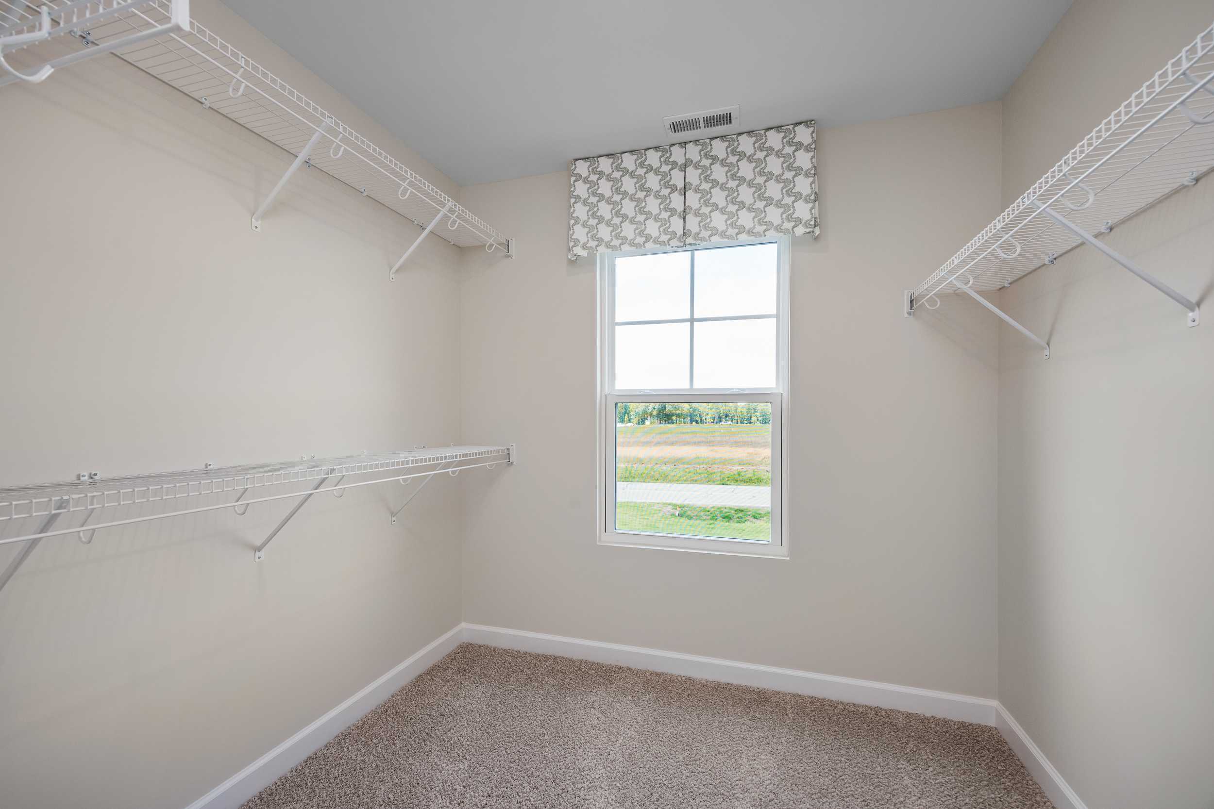 Spacious walk-in closet with white wire shelving, valance window overlooking fields, and carpeted floor at Retreat at North Main in Lillington NC