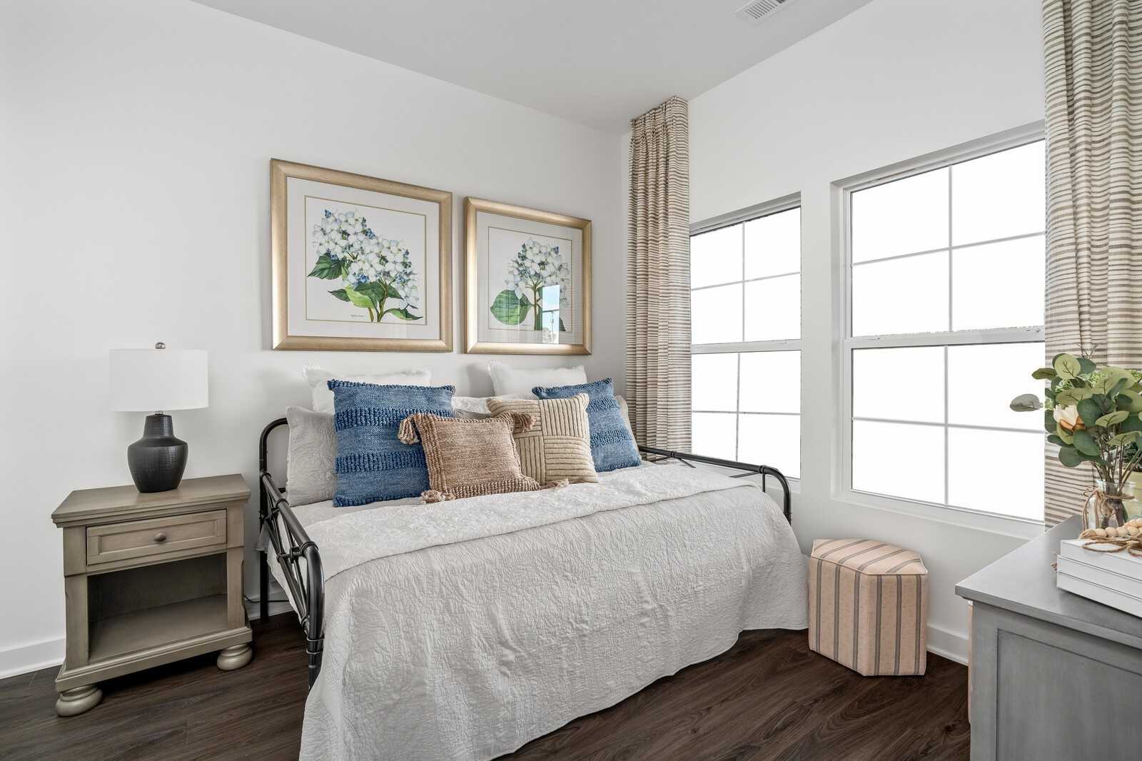 Cozy bedroom with daybed, blue and beige pillows, hydrangea artwork, lamp, and windows in Davidson Homes The Cumberland A Interior, Gallatin, TN