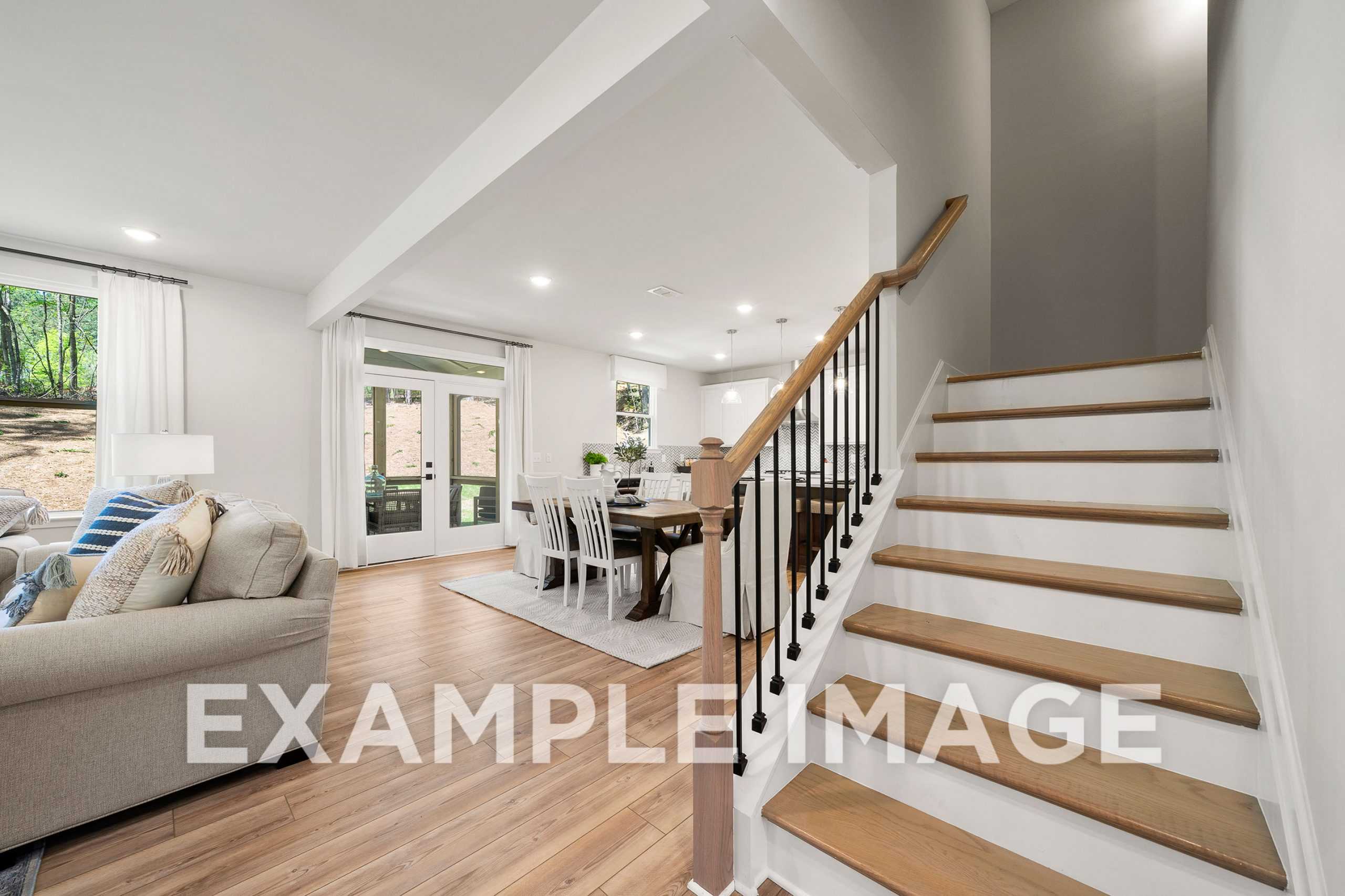 Open-concept living and dining in The Hickory A home, featuring gray sectional sofa, wooden table with white chairs, oak staircase, French doors to greenery