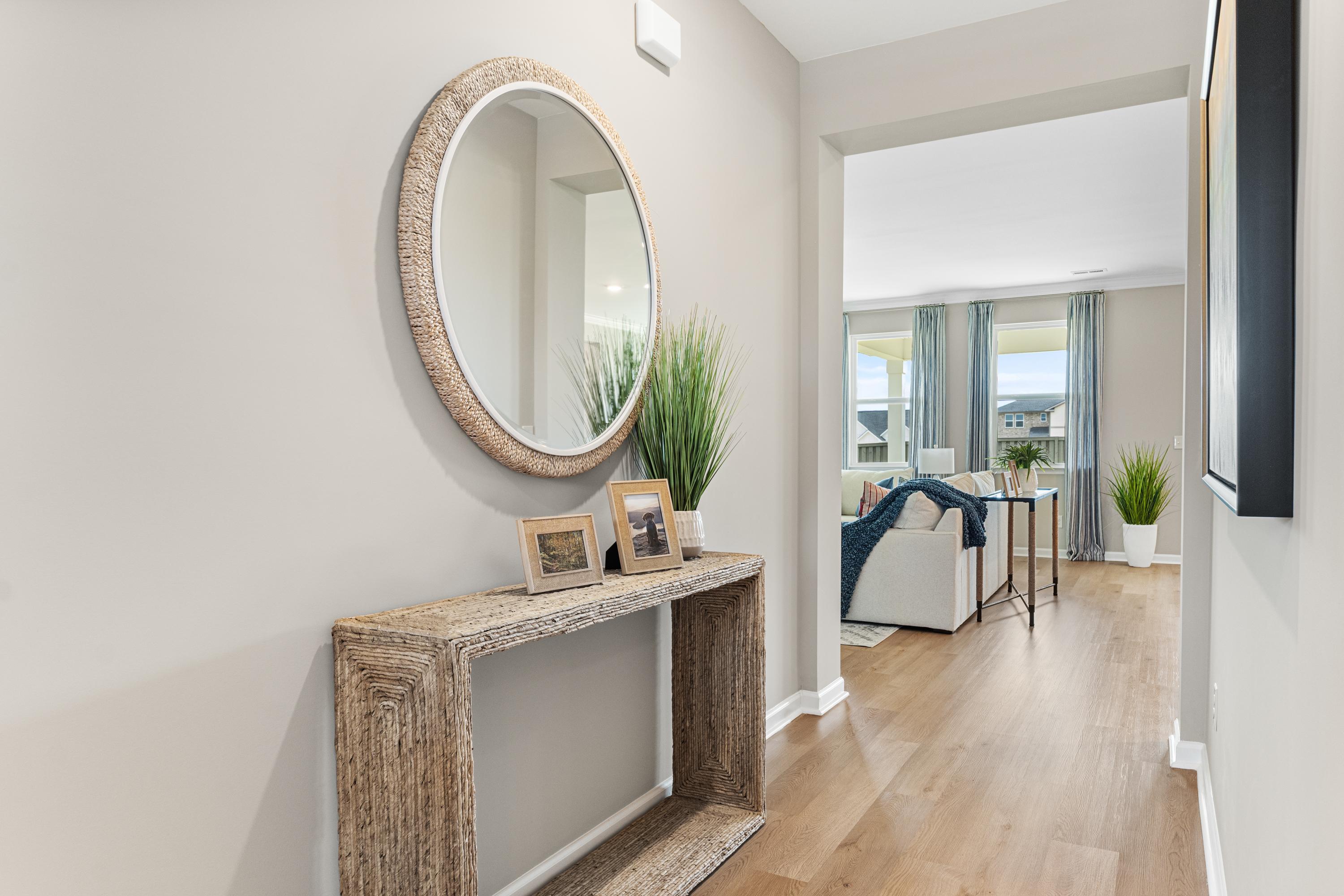 Neutral-toned entryway hallway at The Meadows at Hampton Cove in Owens Cross Roads Alabama with round woven mirror wooden console plants and living room view