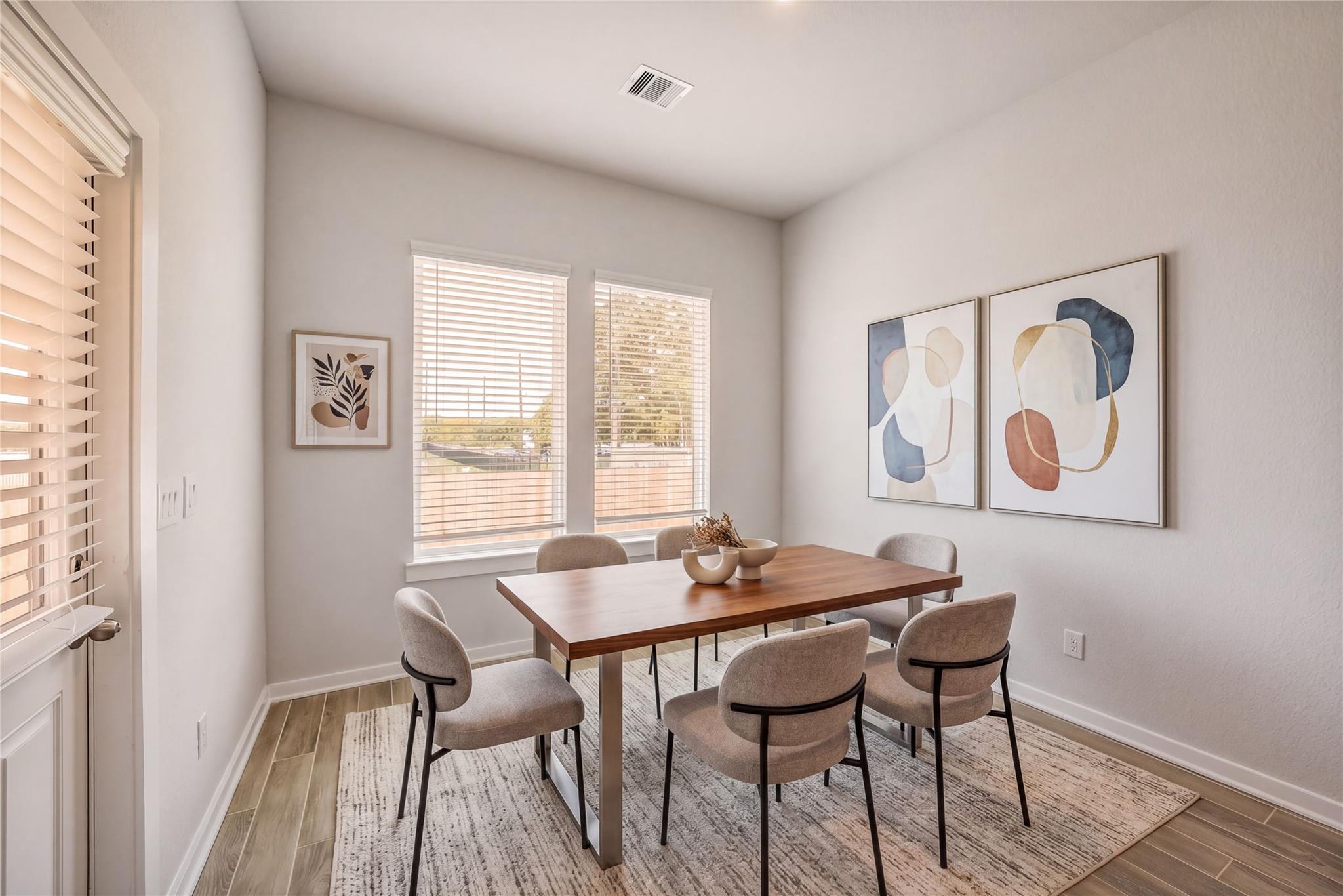 Modern dining room featuring wooden table, beige upholstered chairs, abstract wall art, and window views in The Acadia C by Davidson Homes, Magnolia TX