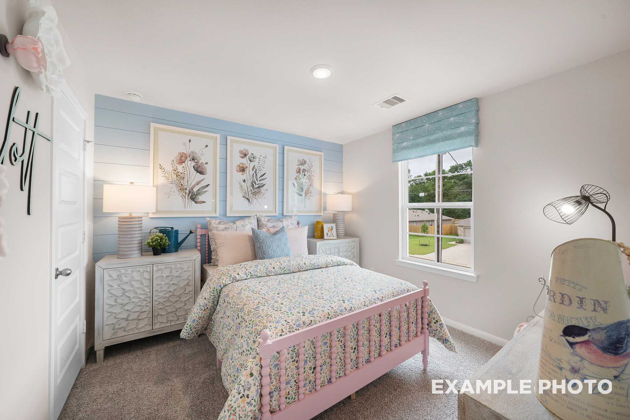 Charming girl's bedroom in The Trinity floor plan with floral pink bed, blue accent wall, artwork, and yard-view window