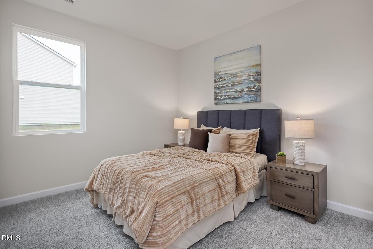 Cozy bedroom with navy upholstered headboard bed, beige textured bedding, wooden nightstands, lamps, abstract art, and large window in Davidson Homes The Carter C, Lillington, NC