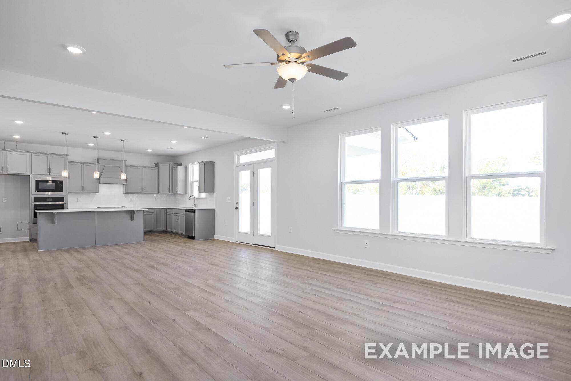 Open-concept kitchen and living area with gray cabinets, island sink, stainless appliances, ceiling fan, and large windows in The Hickory II B Lillington home