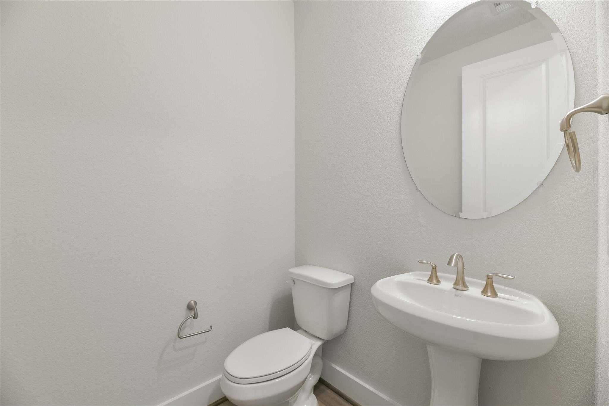 Elegant powder room with round mirror, pedestal sink, gold faucets, and toilet in Davidson Homes The Edward A, Texas City