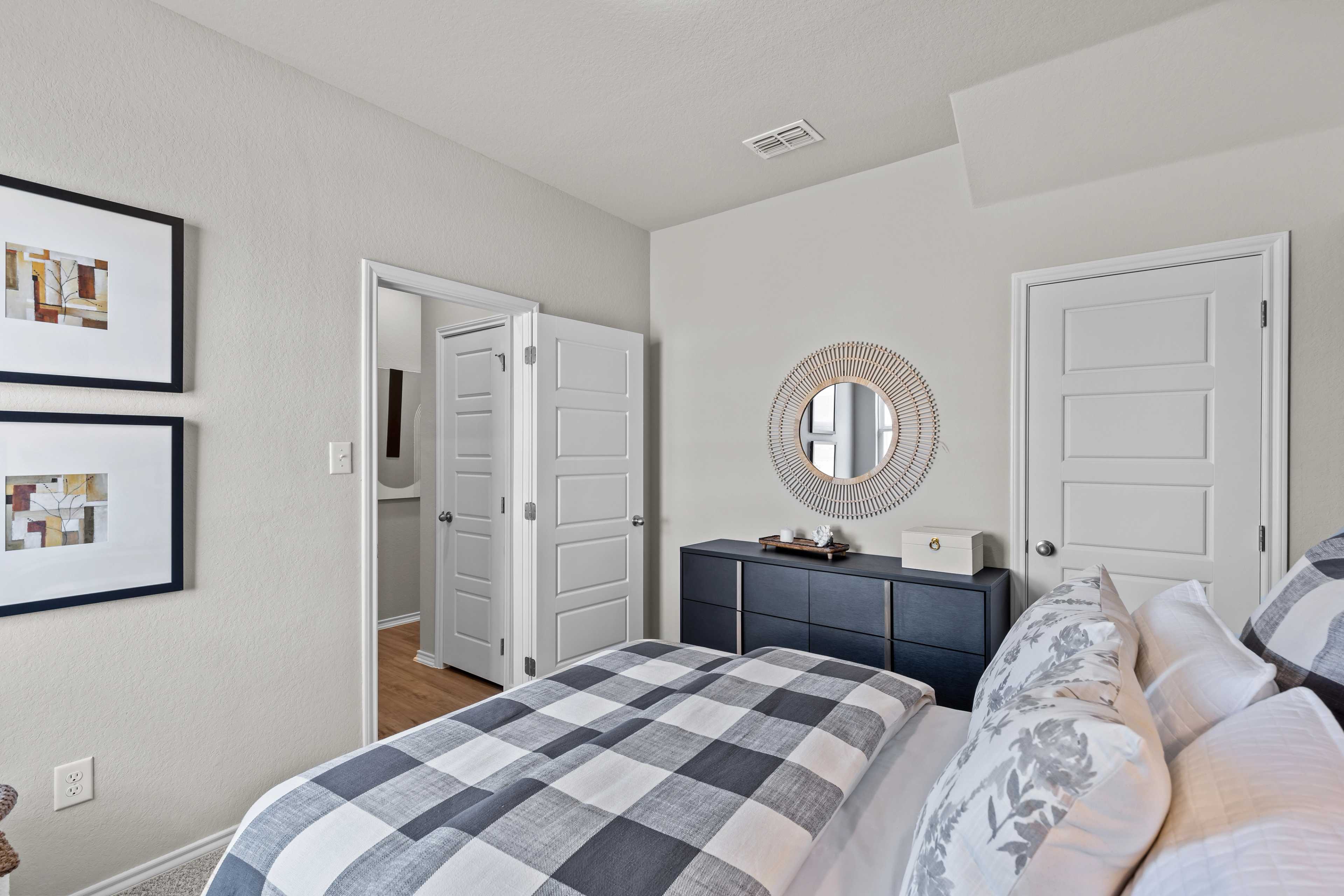 Spacious master bedroom in The Sabine C with checkered duvet, king bed, dresser, round mirror, artwork, and bathroom door