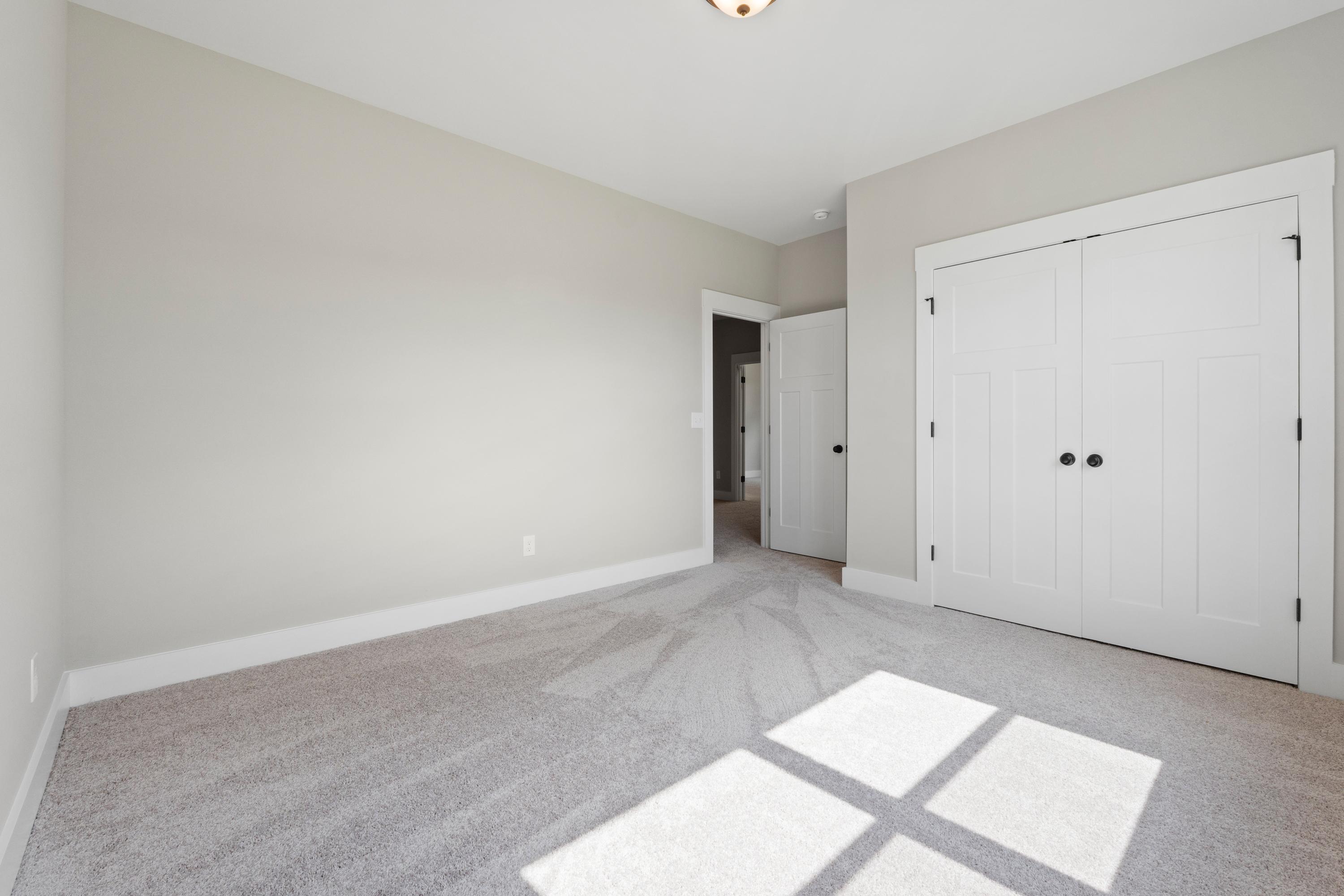 Spacious bedroom interior in The Oxford by Evermore Homes with light gray walls, white double doors, and sunlit carpet floor