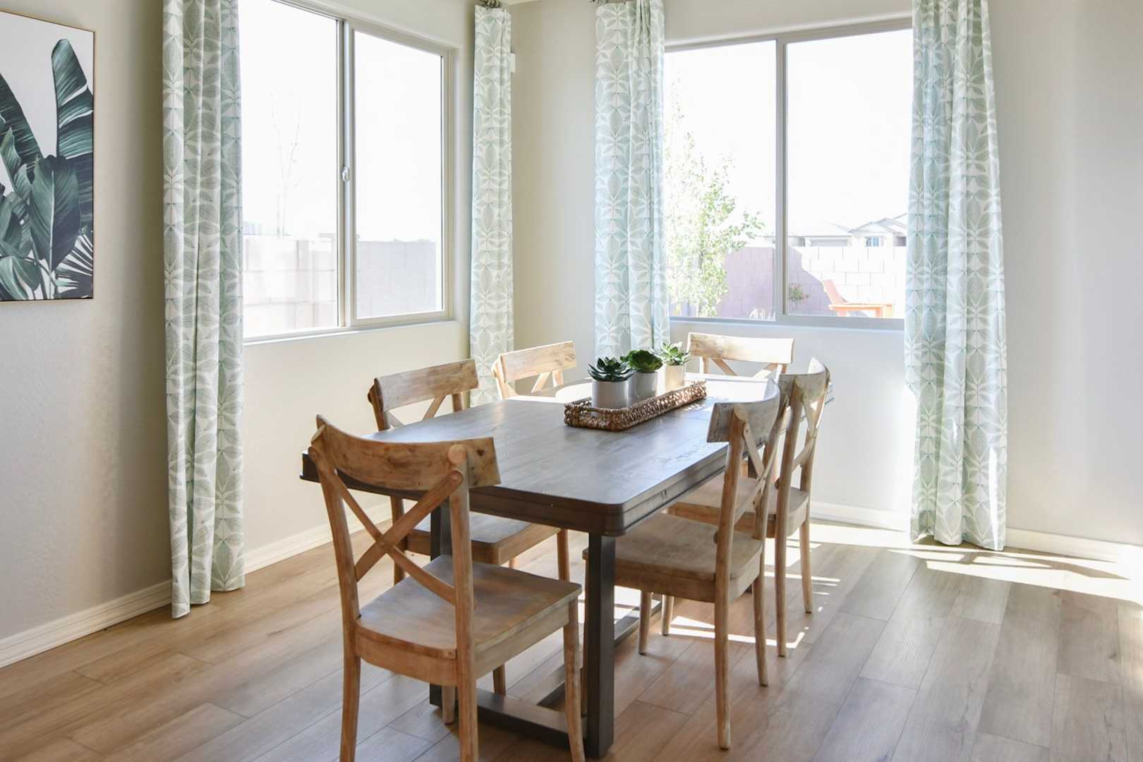 Spacious dining room in The Harmony home design with wooden table, chairs, large windows, sheer curtains, and tropical wall art