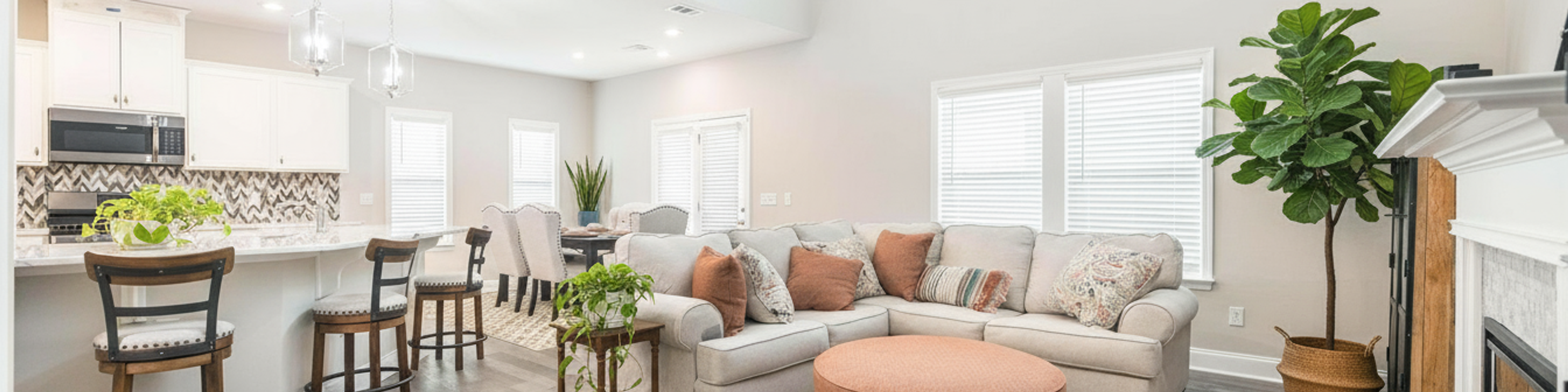 Spacious open-concept kitchen and living room in Links Crossing Auburn Alabama with white cabinets, plush beige sofa, orange ottoman, and fiddle leaf fig plant