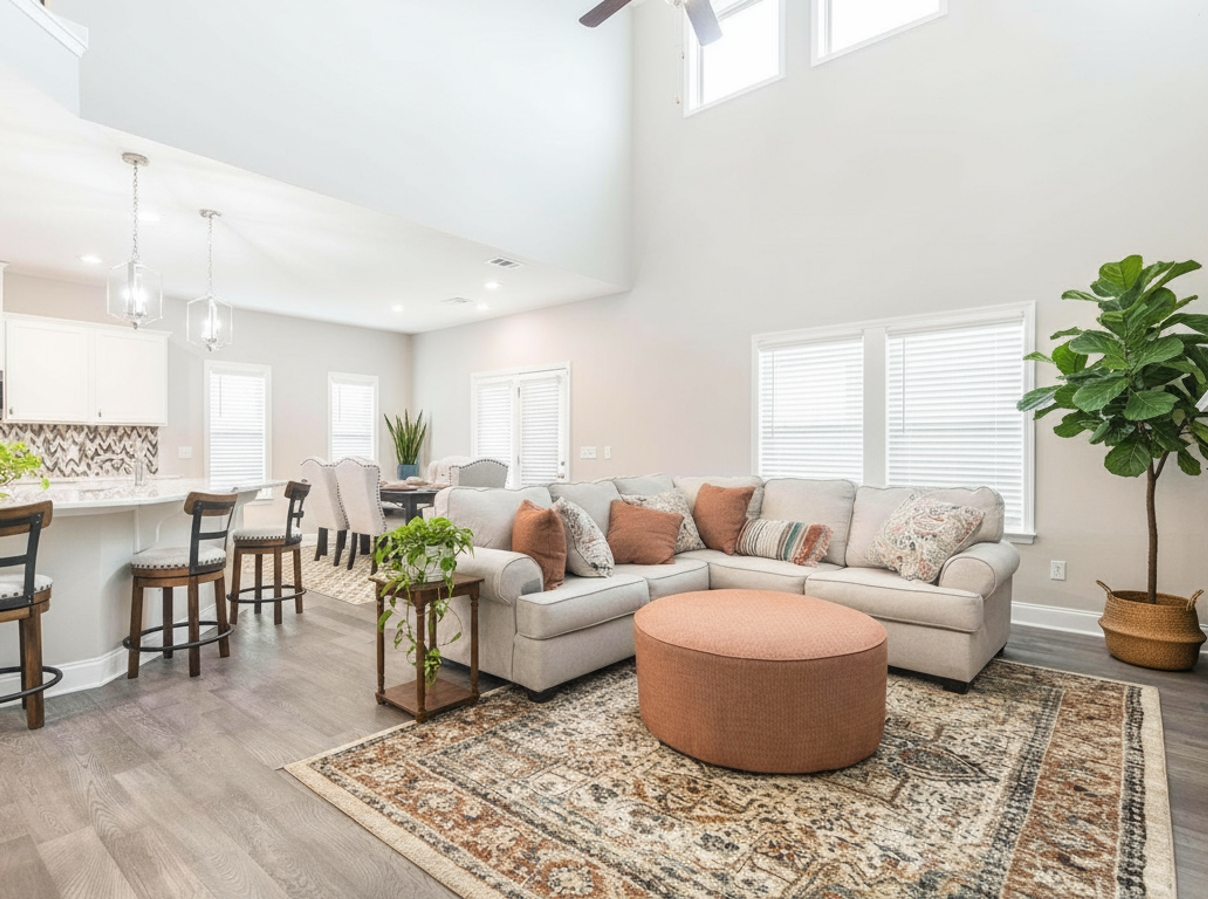 Spacious open-concept kitchen and living room in Links Crossing Auburn Alabama with white cabinets, plush beige sofa, orange ottoman, and fiddle leaf fig plant
