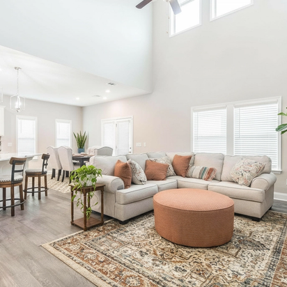 Spacious open-concept kitchen and living room in Links Crossing Auburn Alabama with white cabinets, plush beige sofa, orange ottoman, and fiddle leaf fig plant