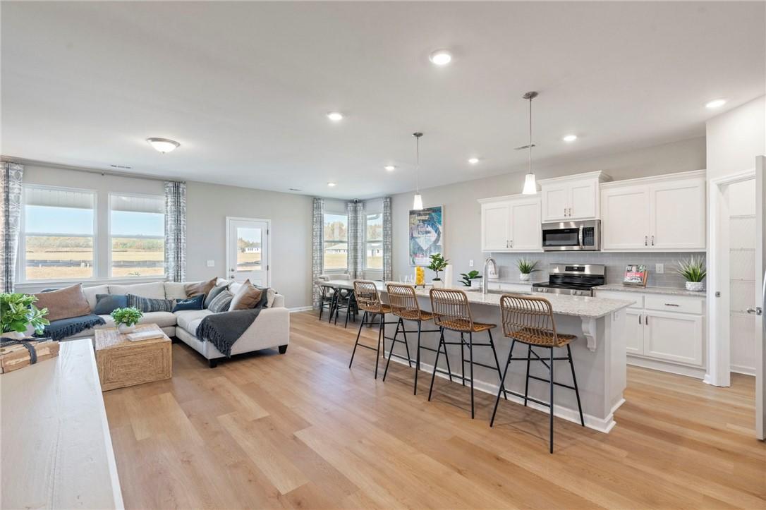 Open-concept kitchen and living room with white cabinets, island bar stools, hardwood floors, and sectional sofa in The Phoenix B, Opelika, Alabama
