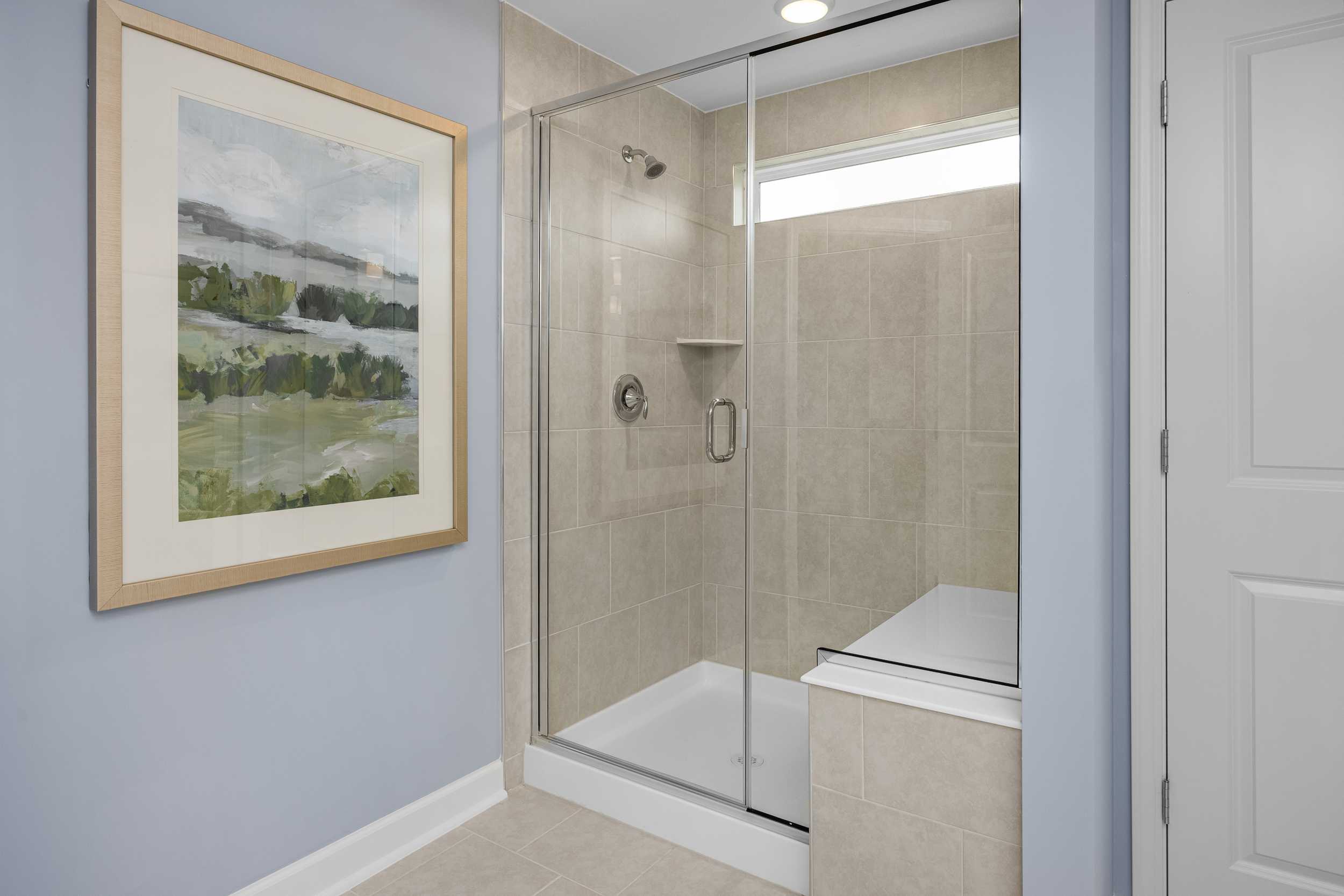 Spacious bathroom at Springvale in Fuquay-Varina NC with frameless glass shower, beige tiles, blue walls, and framed landscape art