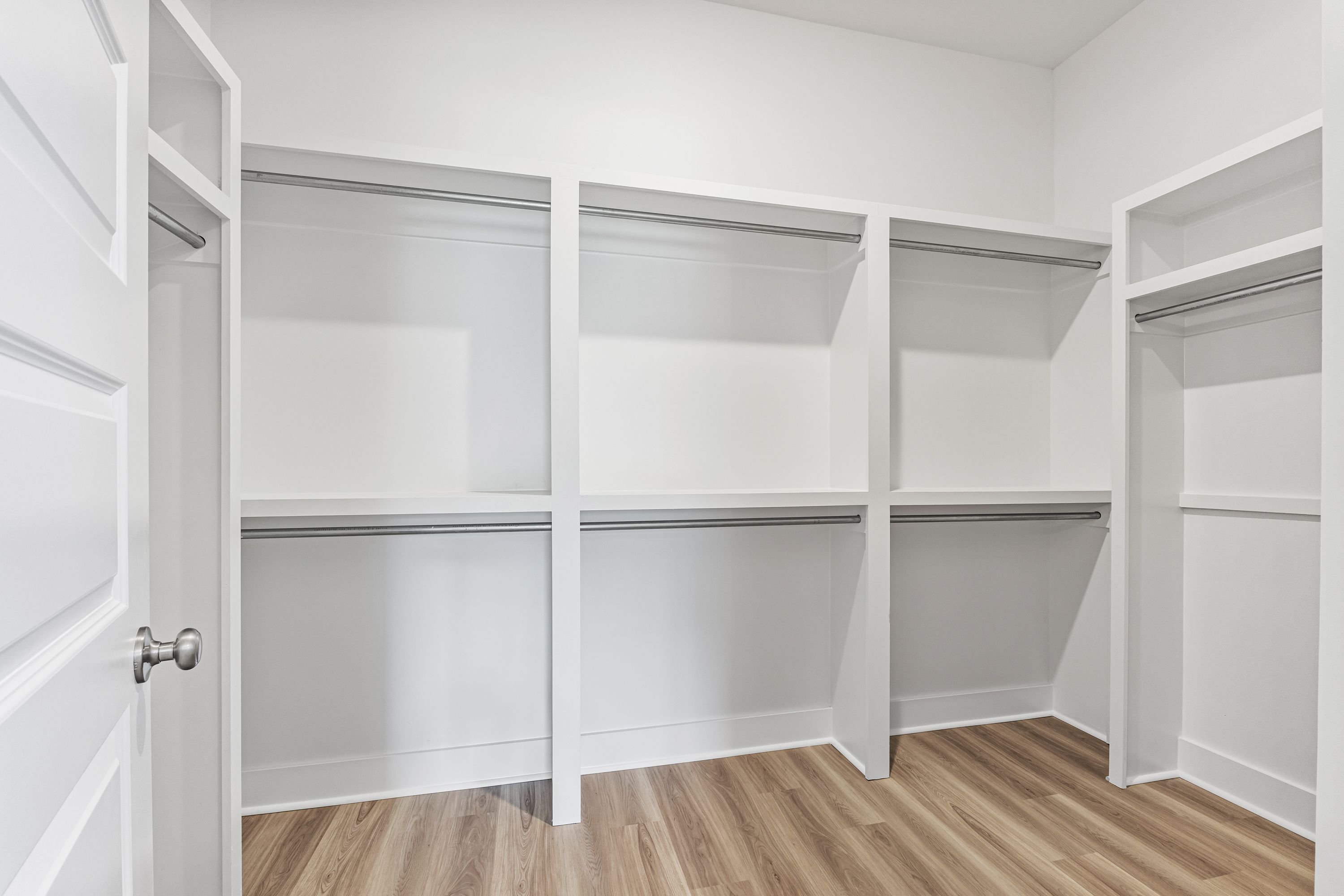 Spacious walk-in closet in The Oxford A master suite with white walls, wood flooring, built-in shelves and hanging rods