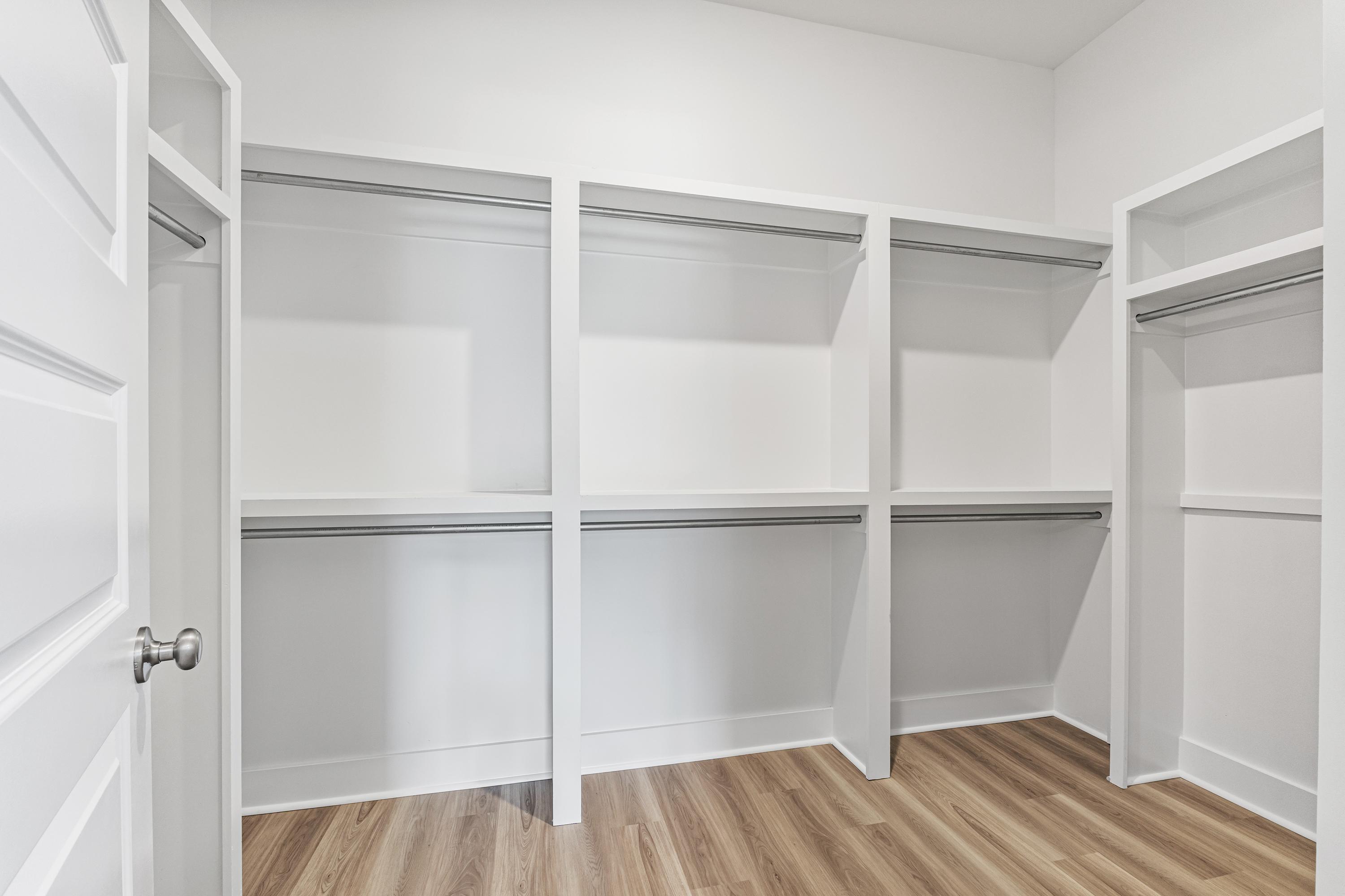 Spacious walk-in closet in The Oxford A master suite with white walls, wood flooring, built-in shelves and hanging rods