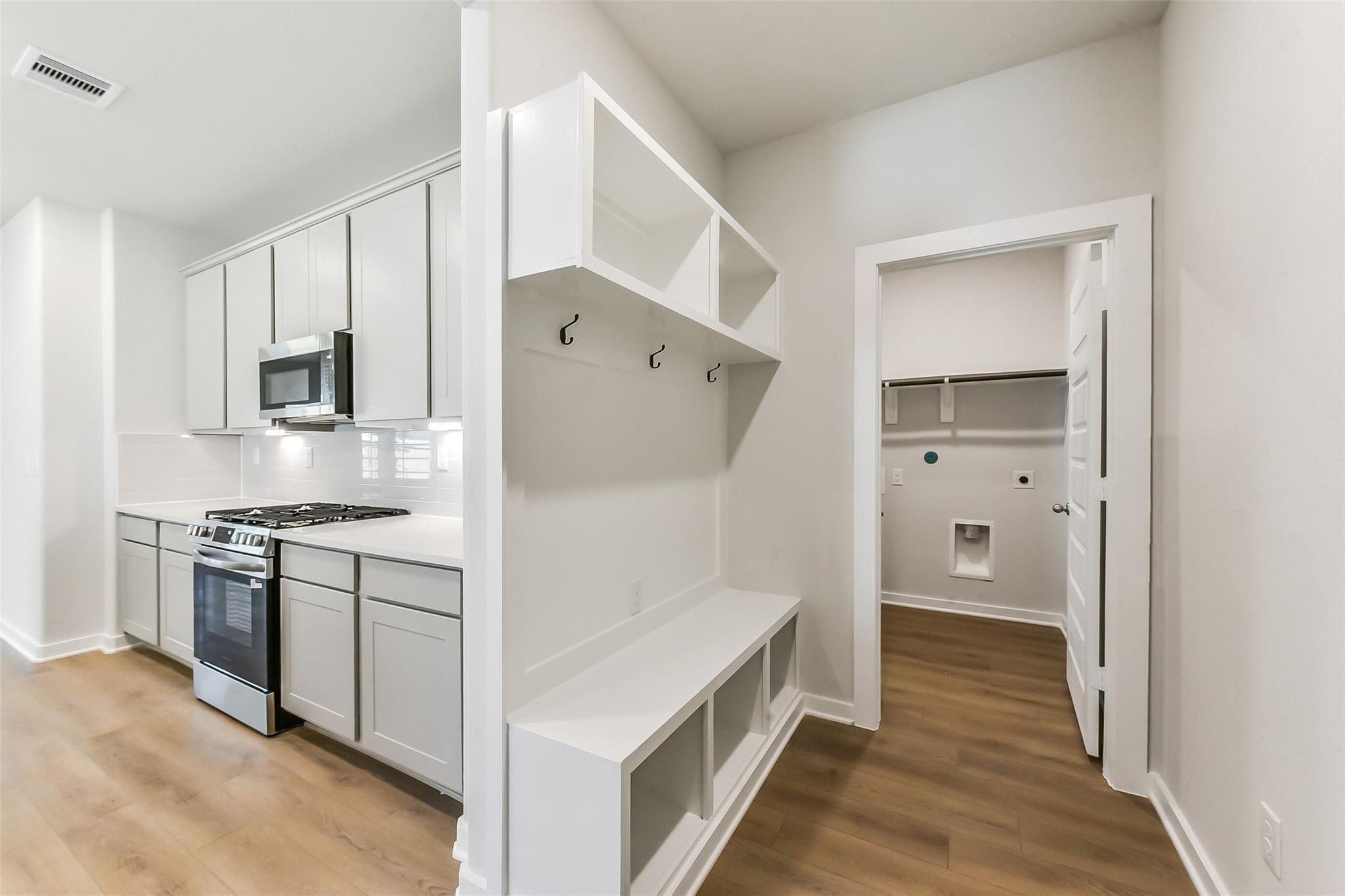 Modern white kitchen with stainless appliances adjacent to mudroom bench, hooks, and laundry in Davidson Homes The Brazos E, Magnolia TX