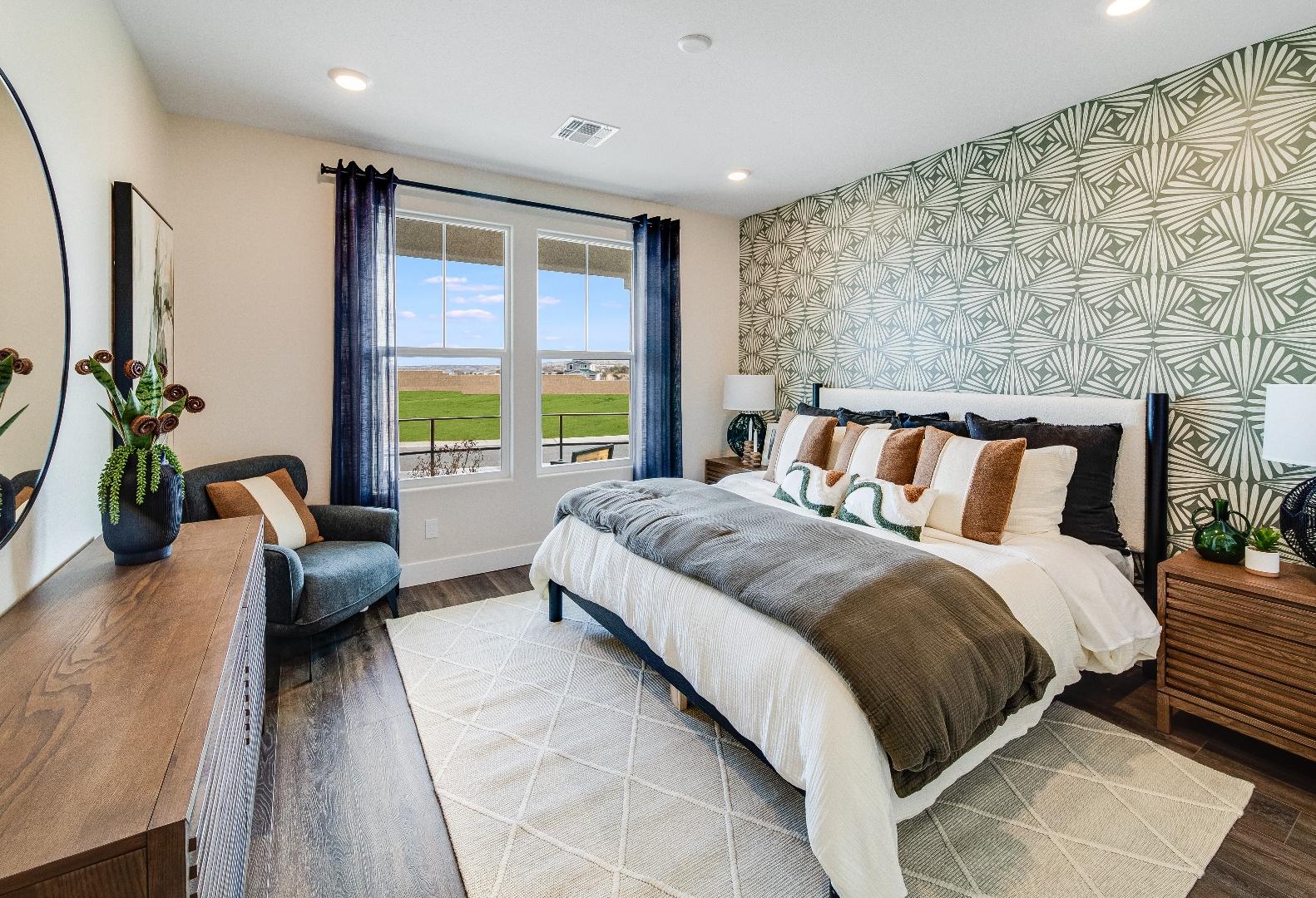 Spacious master bedroom in The Boulder II with king bed, geometric green wallpaper, large window view of fields, wooden nightstands