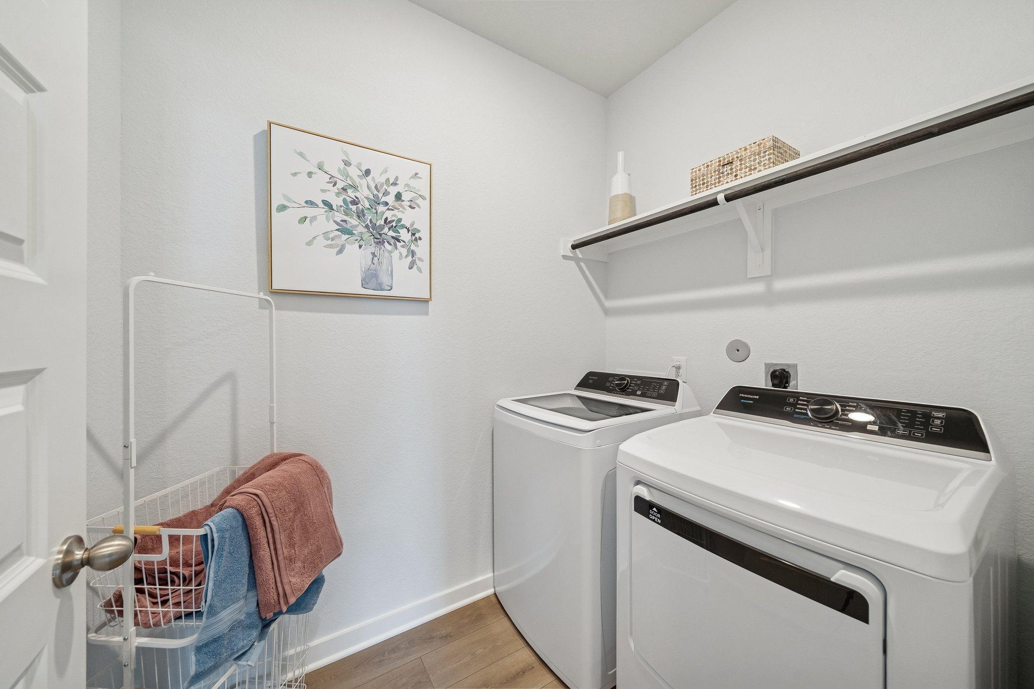 Spacious laundry room in Spring Branch Crossing Conroe TX with white washer dryer, wooden shelves, floral art, and ironing board