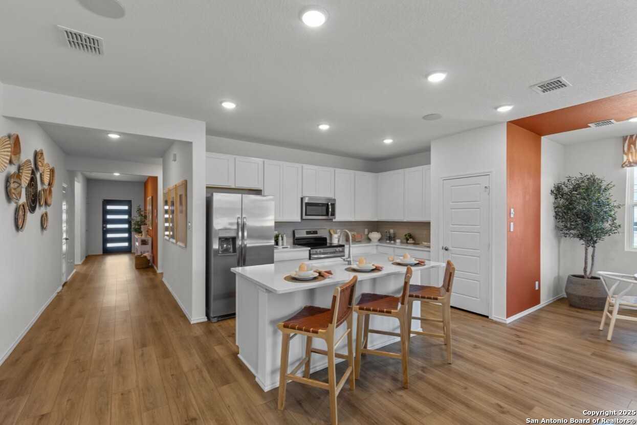 Modern open-concept kitchen with white cabinets, stainless appliances, island bar stools, and hallway in Davidson Homes The Daphne E, Elmendorf, Texas