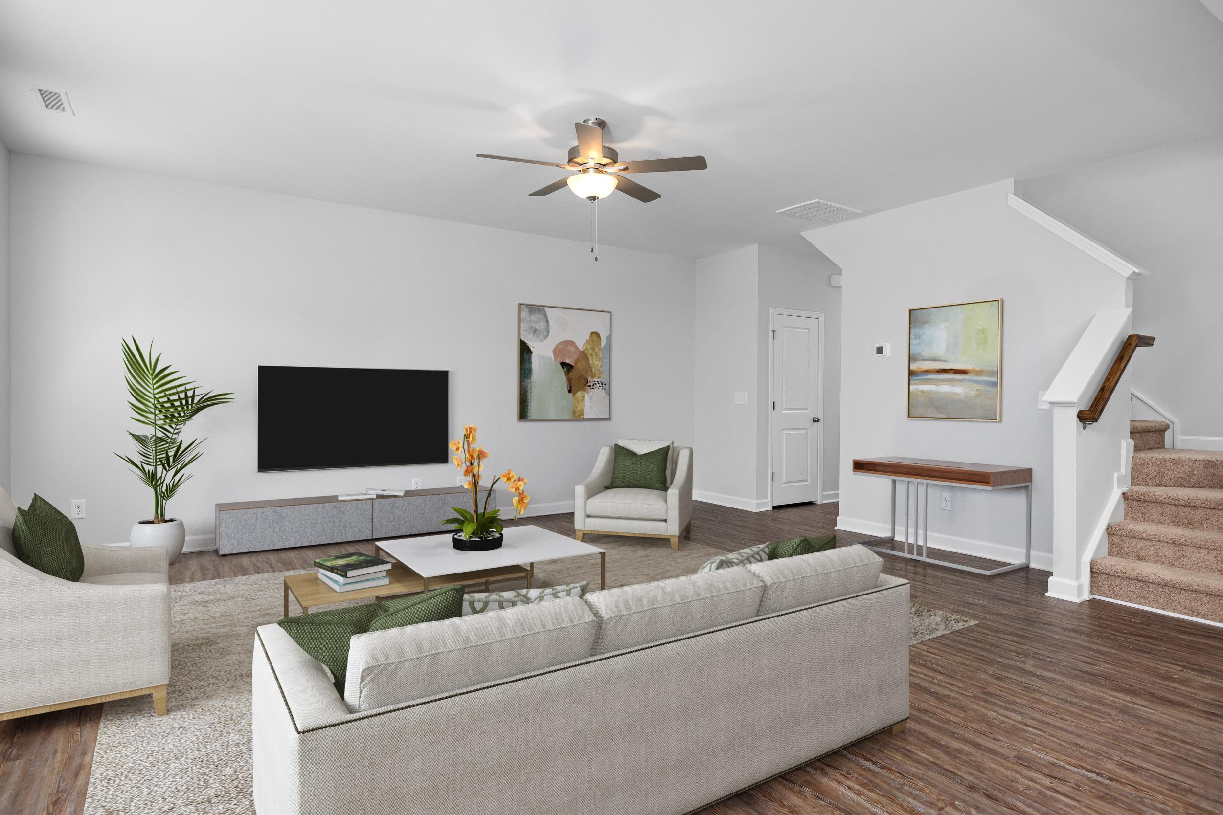 Spacious living room in Gregory Village Townhomes, Lillington NC featuring white sofa, wooden floors, TV, plants, and staircase by Davidson Homes