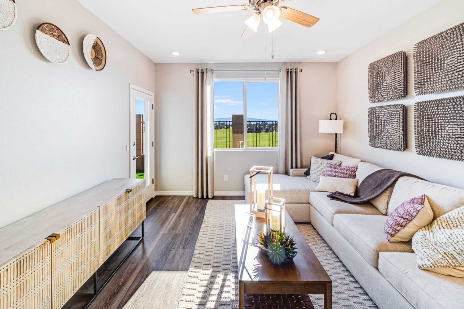 Spacious living room in The Savannah C with L-shaped beige sofa, woven wall art, wooden console, and scenic window views of Prescott fields