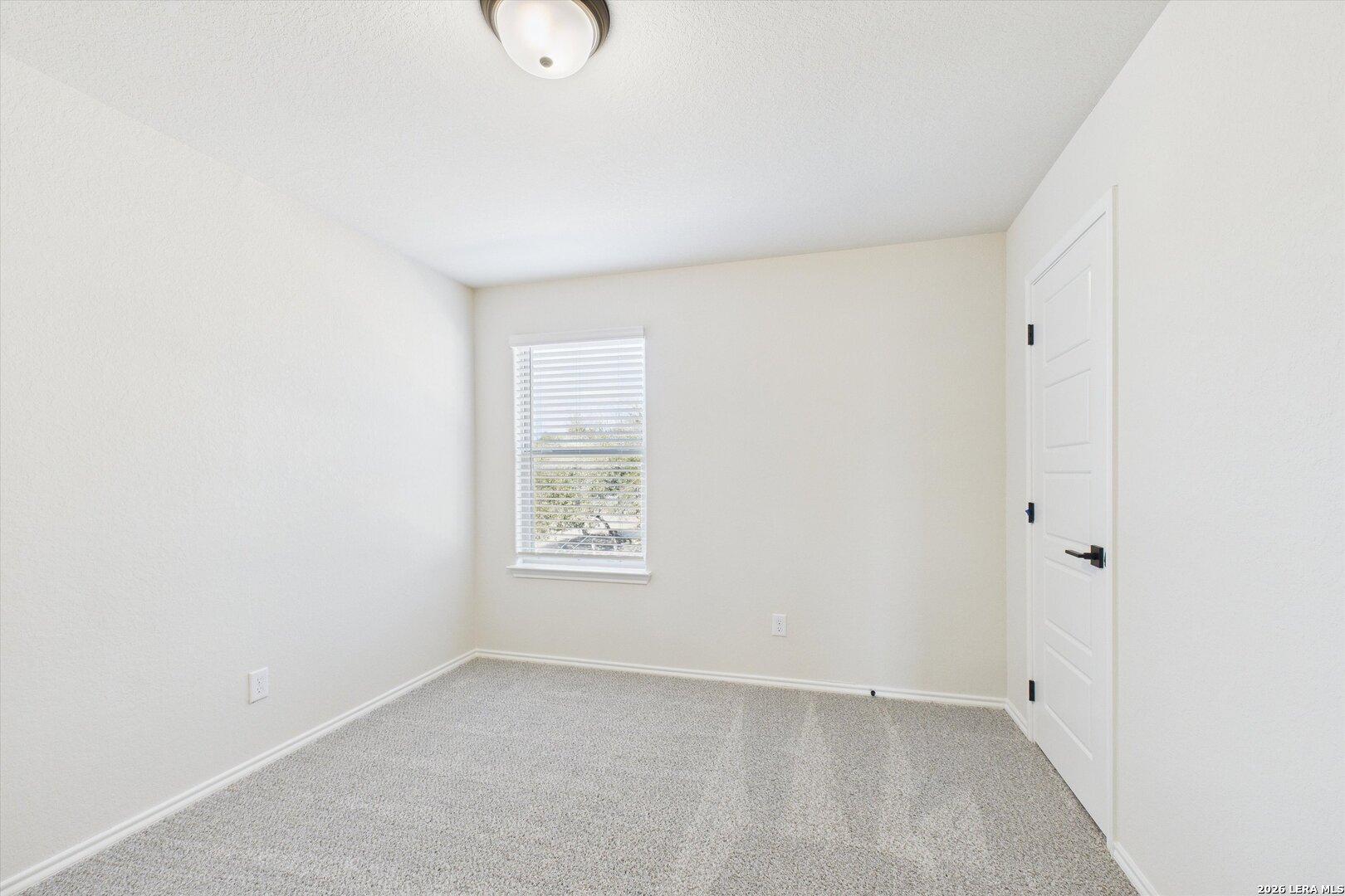 Spacious empty bedroom with beige walls, carpet flooring, window blinds and door in Davidson Homes The Gillian B, San Antonio