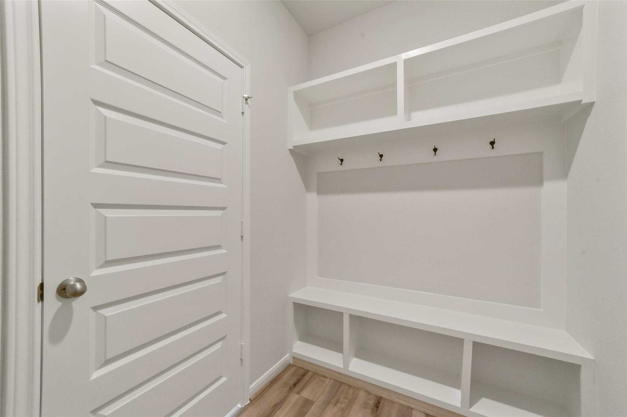 White mudroom with built-in bench, open shelves, and coat hooks beside paneled door in Davidson Homes San Marcos E, Beasley Texas