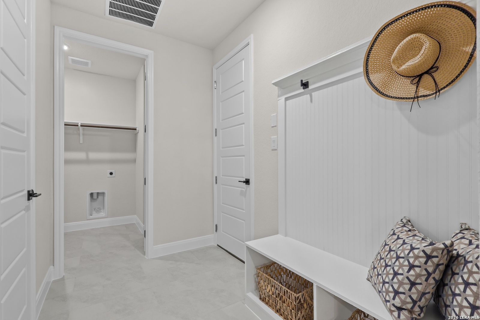 Functional mudroom with built-in bench, patterned pillows, wicker basket, and adjacent laundry in Davidson Homes The Lanier G, Castroville, Texas