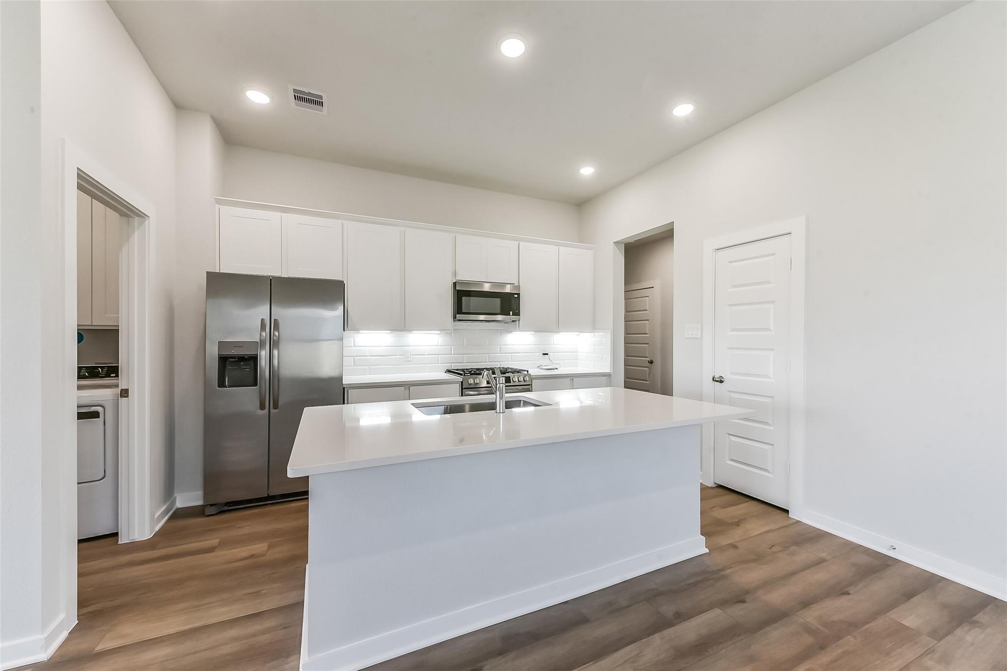Modern white kitchen with quartz island, stainless steel appliances, subway tile backsplash, and hardwood floors in Davidson Homes The Costa C, Dayton, Texas