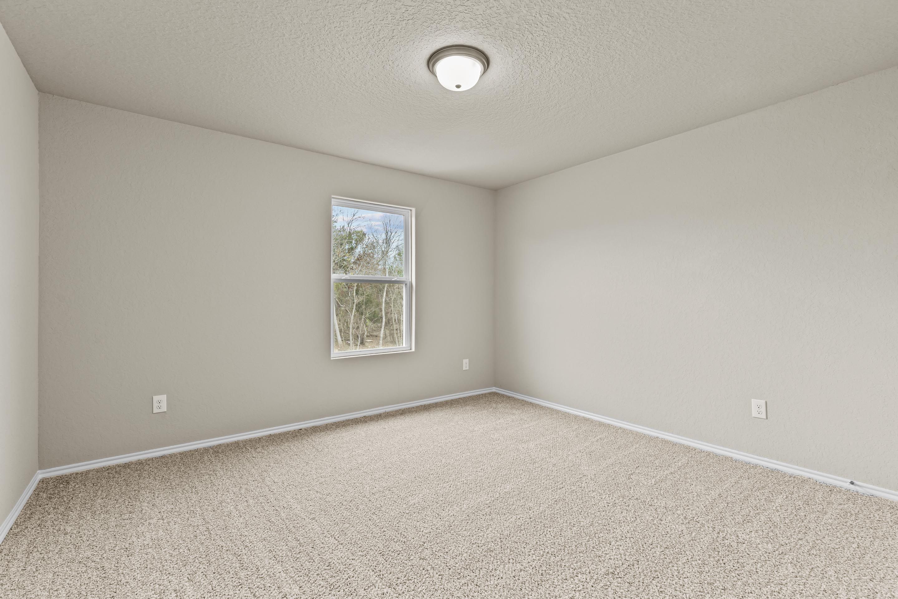 Bright empty bedroom with light gray walls, beige carpet, large window with blinds, and flush-mount ceiling light in Davidson Homes The Douglas F, San Antonio