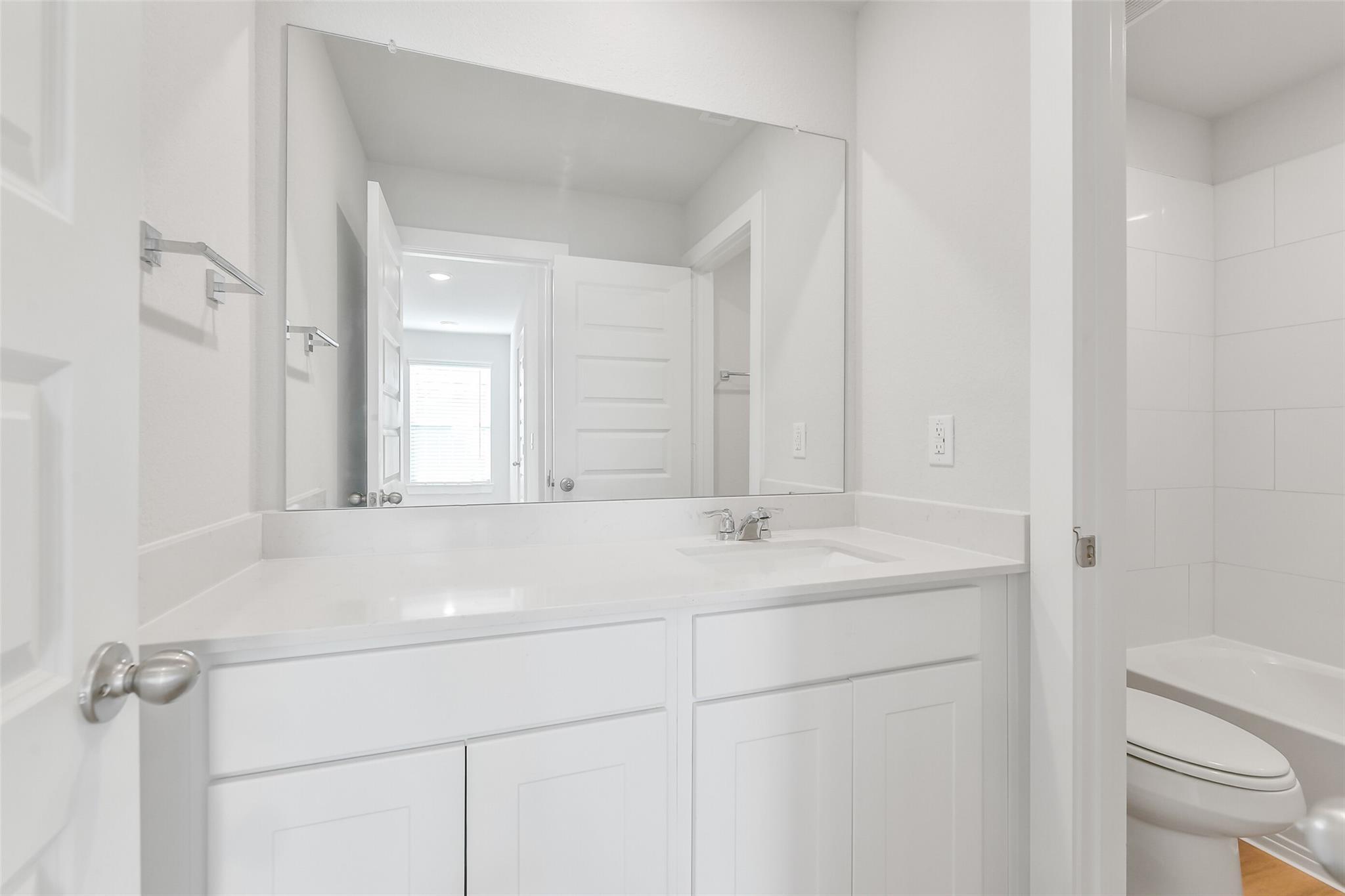 Bright white secondary bathroom with single vanity, large mirror, and toilet in Davidson Homes The Trinity F, Magnolia, Texas
