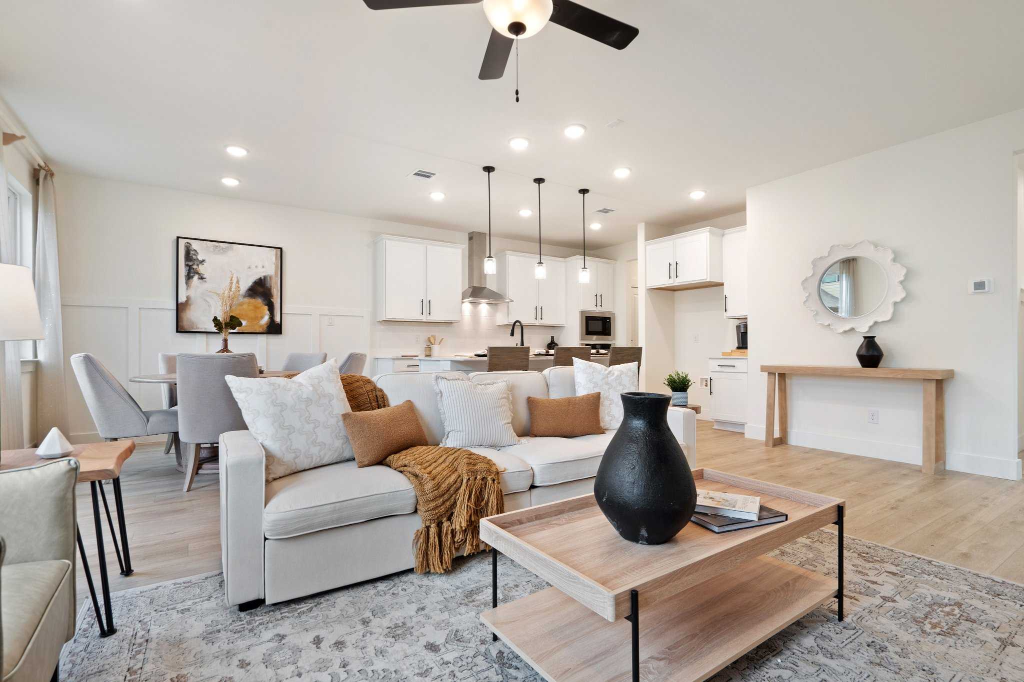 Open-concept living room and kitchen in The Wake: white sofa with throws, wooden coffee table, dining area, pendant lights, beige walls, hardwood floors