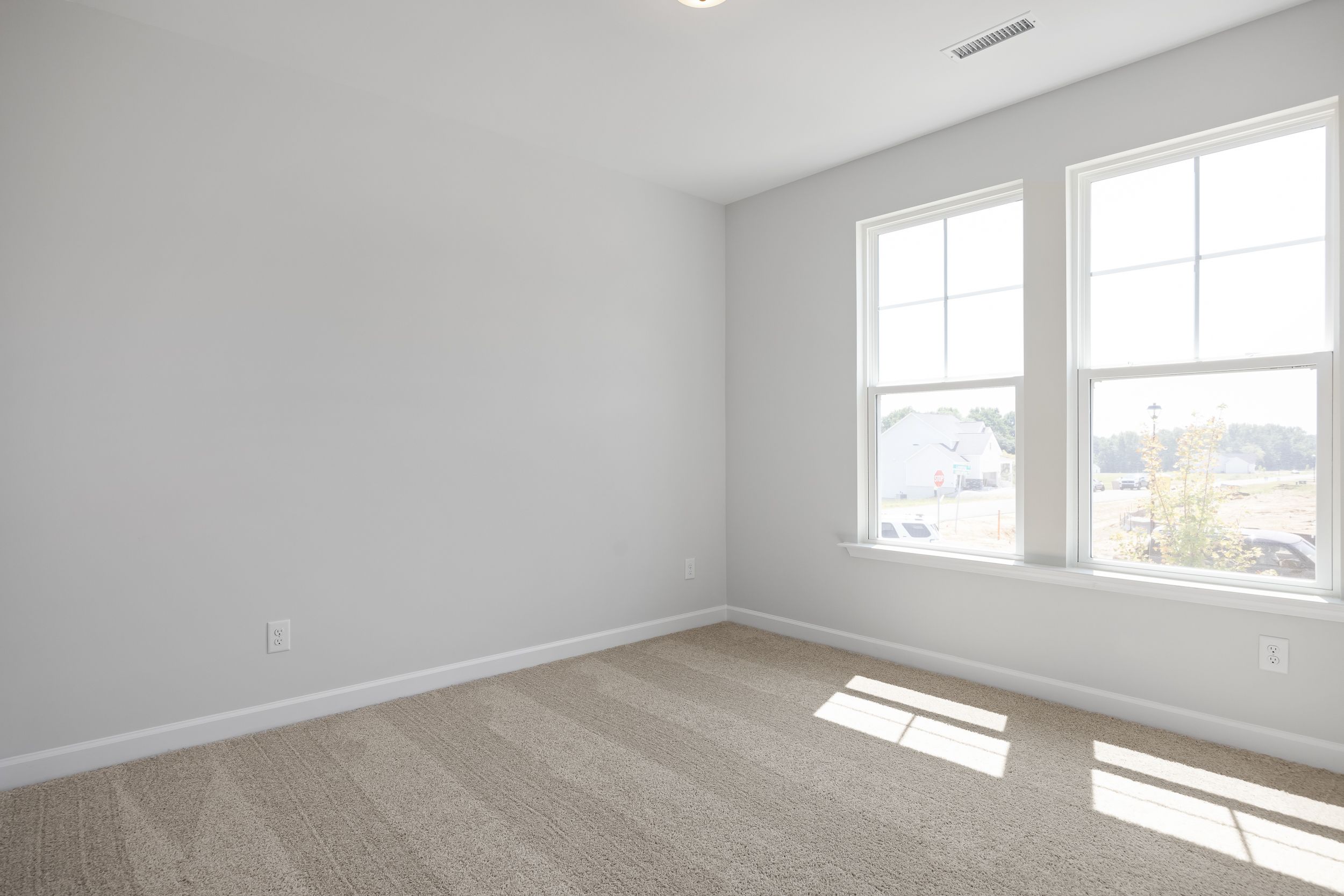 Bright empty bedroom in The Daphne C with gray walls, beige carpet, large sunny windows overlooking suburban view
