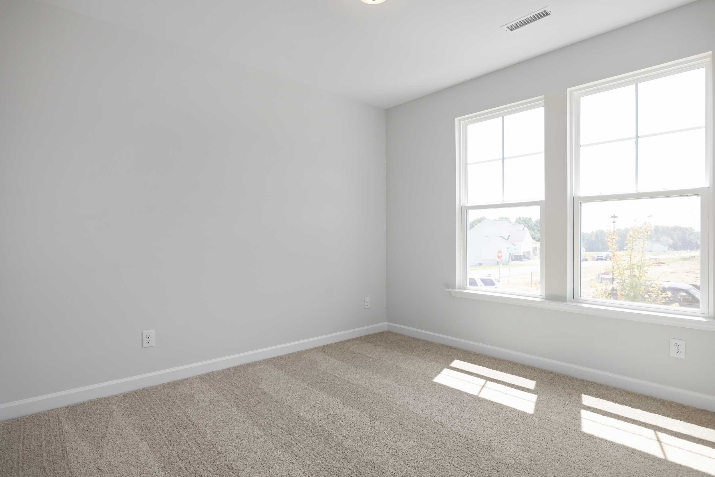 Bright empty bedroom in The Daphne C with gray walls, beige carpet, large sunny windows overlooking suburban view