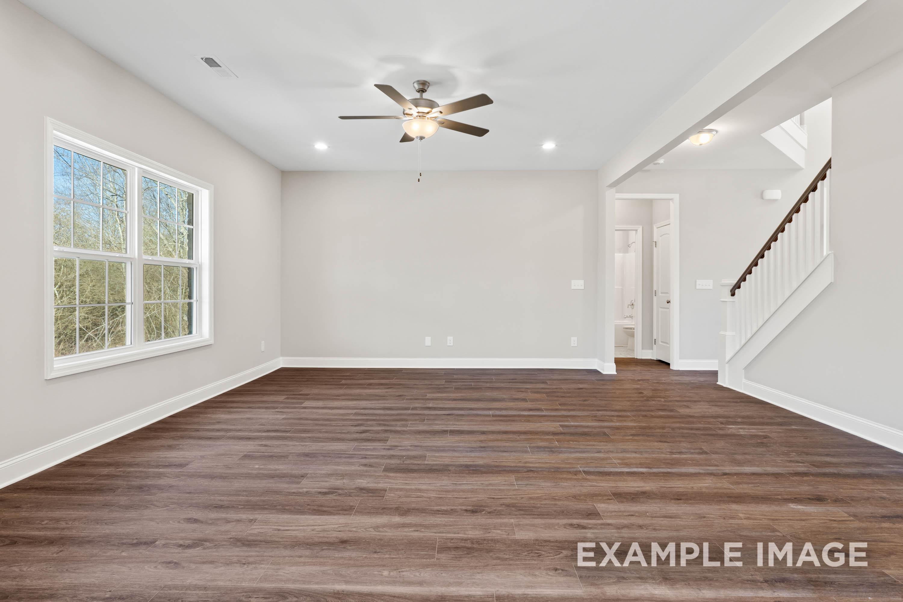 Spacious living room in The Madison B with light gray walls, dark hardwood floors, ceiling fan, large windows, and staircase