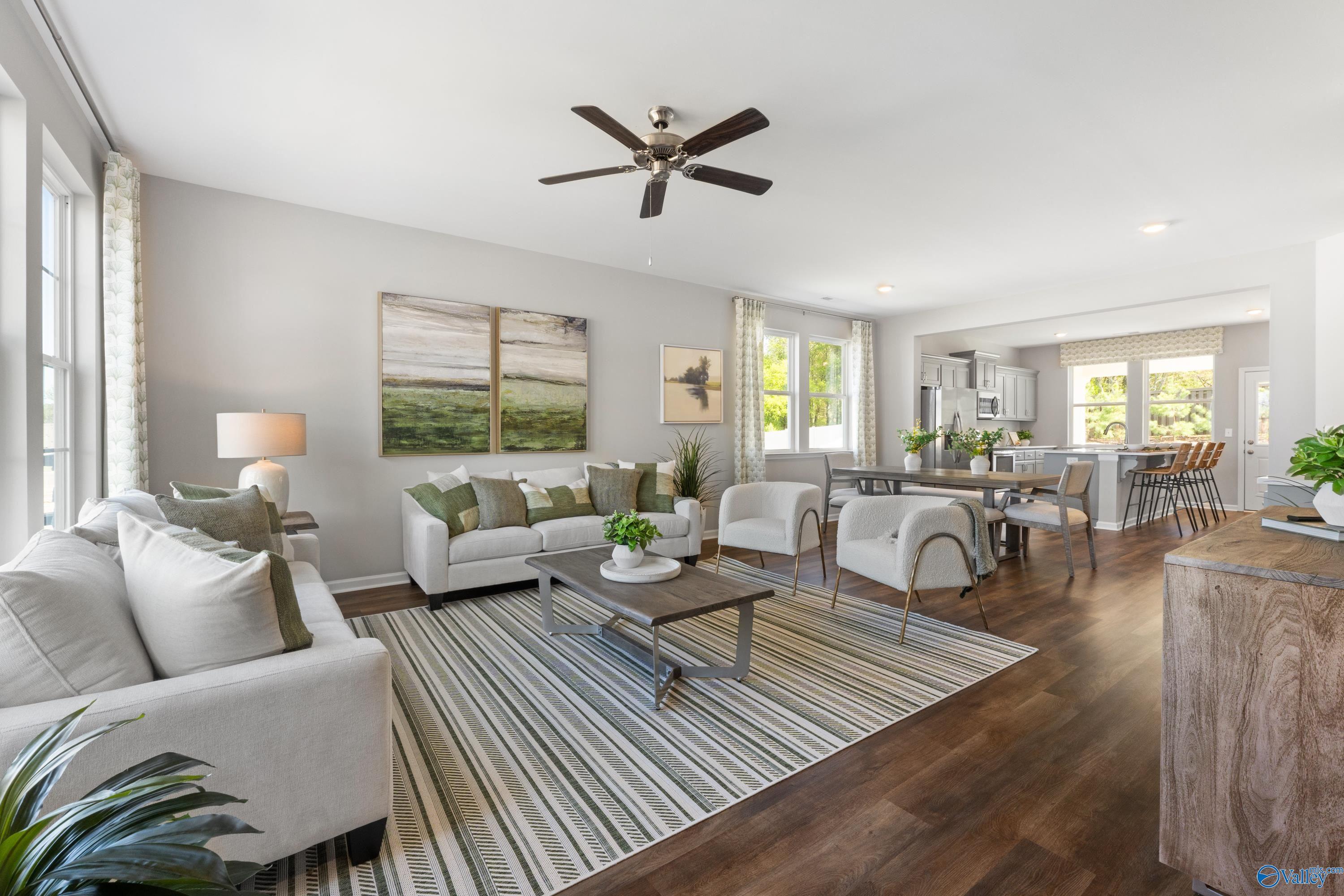 Open-concept living room with white sofas, green accents, ceiling fan, and kitchen views in Davidson Homes The Stella, Madison, Alabama
