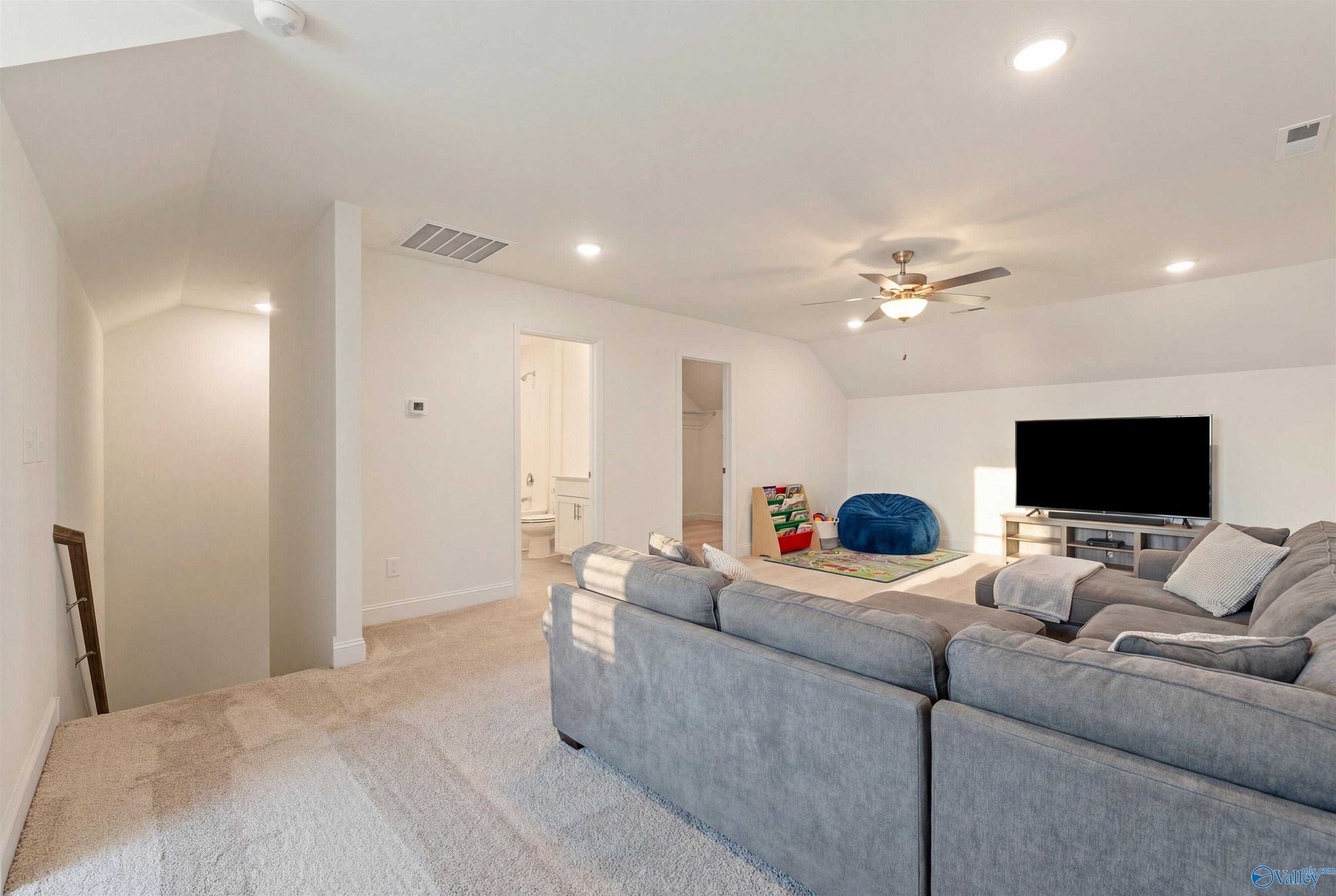 Spacious bonus room with vaulted ceiling, ceiling fan, gray L-shaped sofa, wall-mounted TV, bean bag, and bathroom access in Davidson Homes The Rockford B, Toney, AL