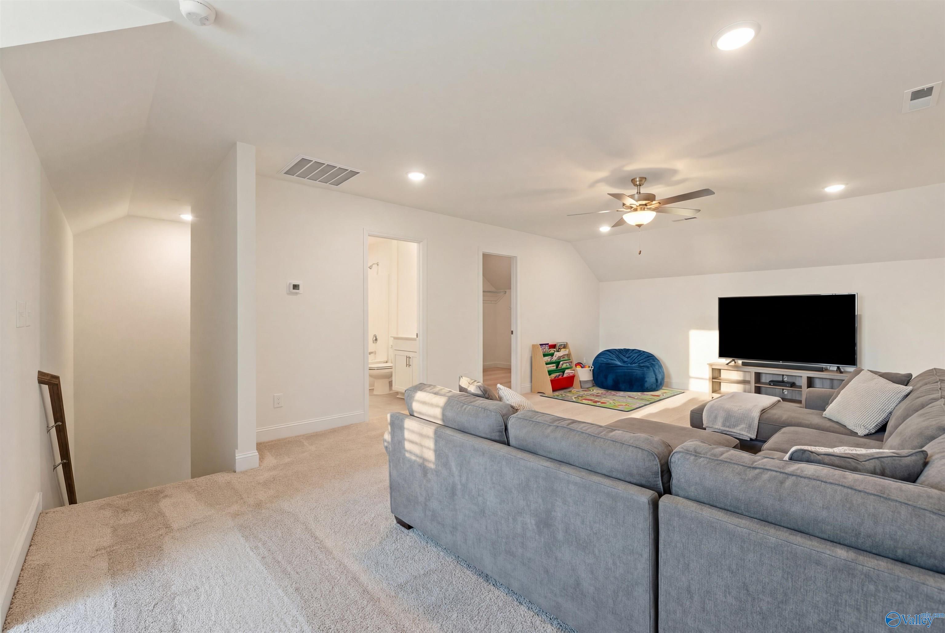 Spacious bonus room with vaulted ceiling, ceiling fan, gray L-shaped sofa, wall-mounted TV, bean bag, and bathroom access in Davidson Homes The Rockford B, Toney, AL