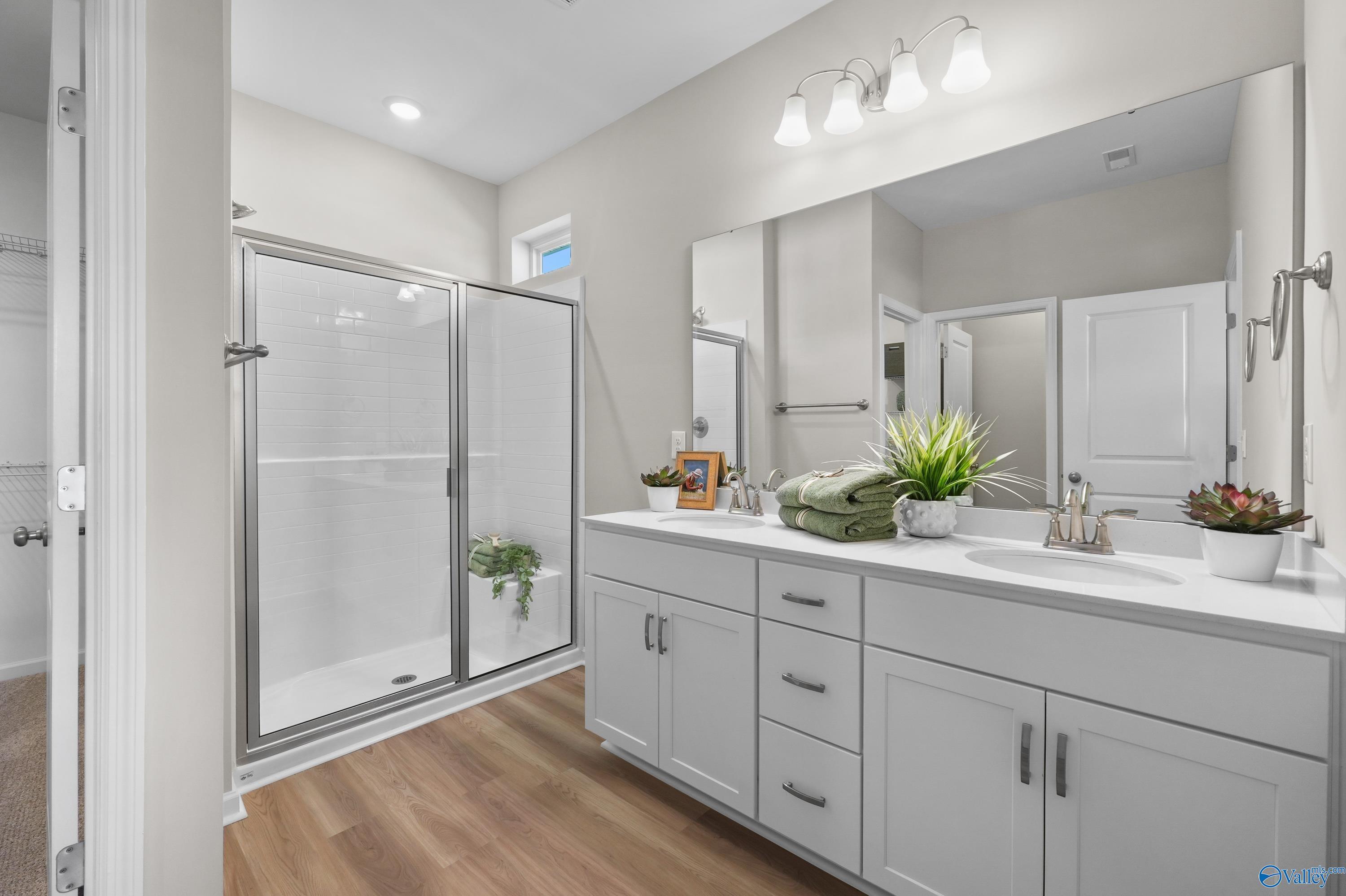 Modern master bathroom with double white vanity, glass walk-in shower, plants, and wood flooring in Evermore Homes The Malibu, Madison, AL