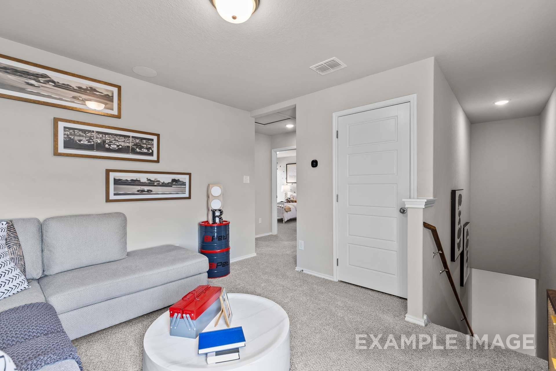 Cozy living room with gray sectional sofa, automotive artwork, red toolbox, and open bedroom view in Davidson Homes Sabine J, Heartland Texas