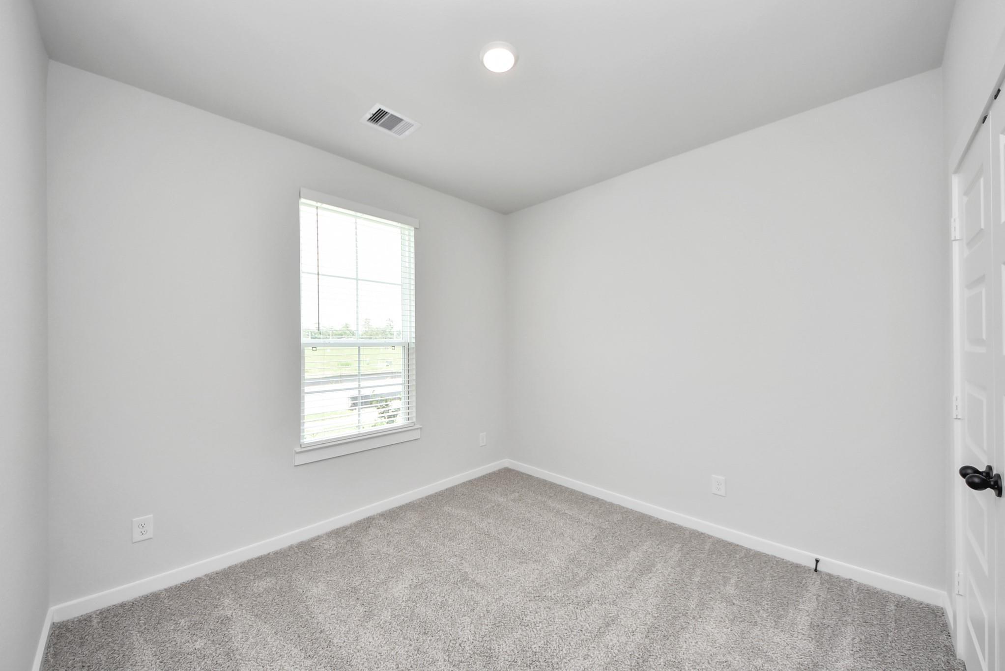Bright empty bedroom with gray carpet, white walls, and window with blinds in Davidson Homes Brookside F plan, Crosby, Texas