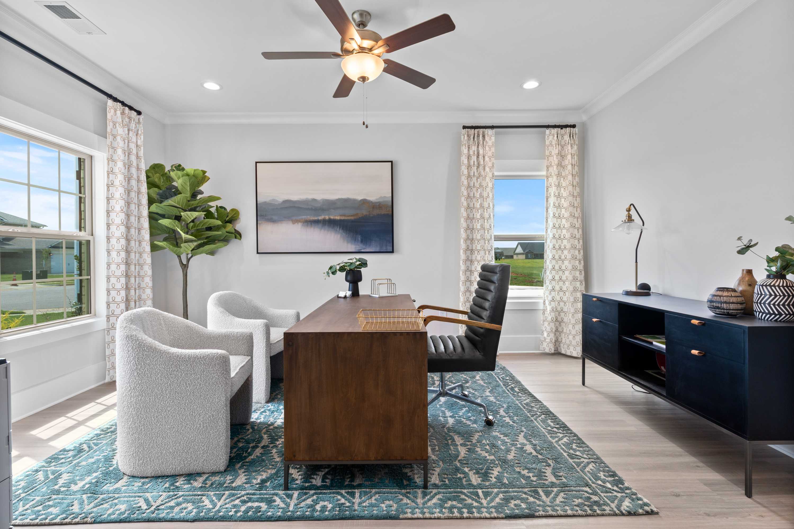 Spacious home office in The Chelsea B with wooden desk, black leather chair, white armchairs, blue rug, and large windows
