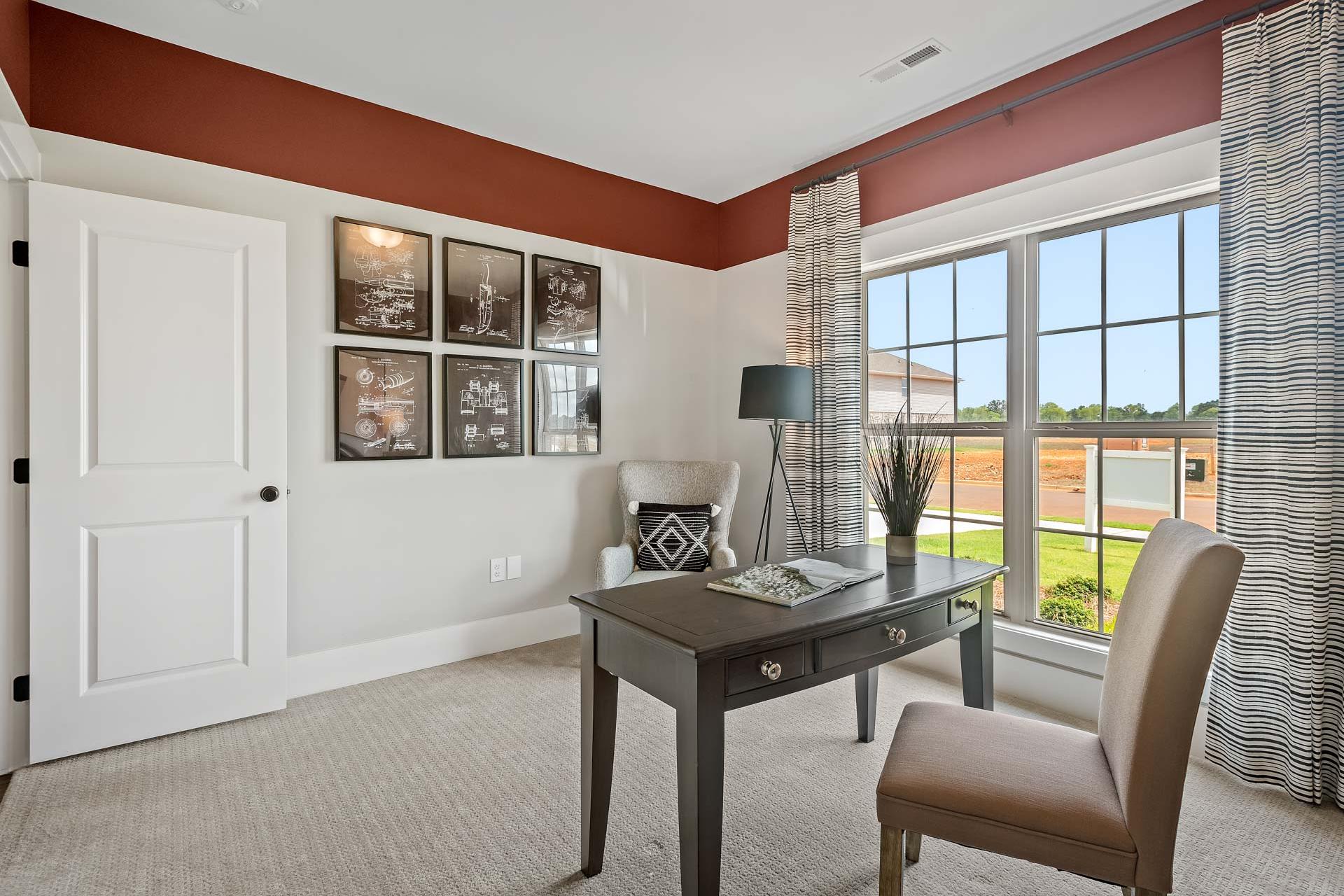 Cozy home office at Heritage Lakes in New Market Alabama with dark wood desk, beige armchair, framed art, and large window views