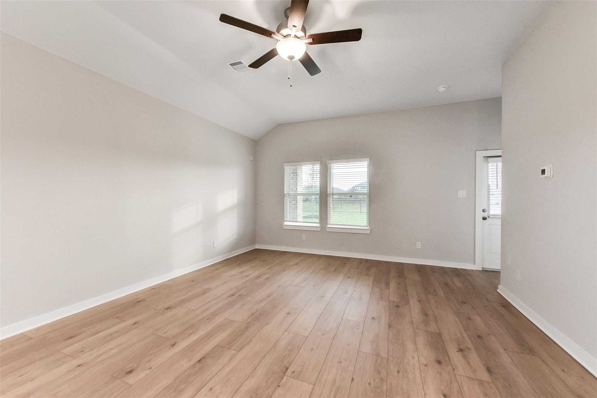 Spacious empty living room with light oak hardwood floors, ceiling fan, and windows to green field in Davidson Homes The Laguna B, Katy, Texas