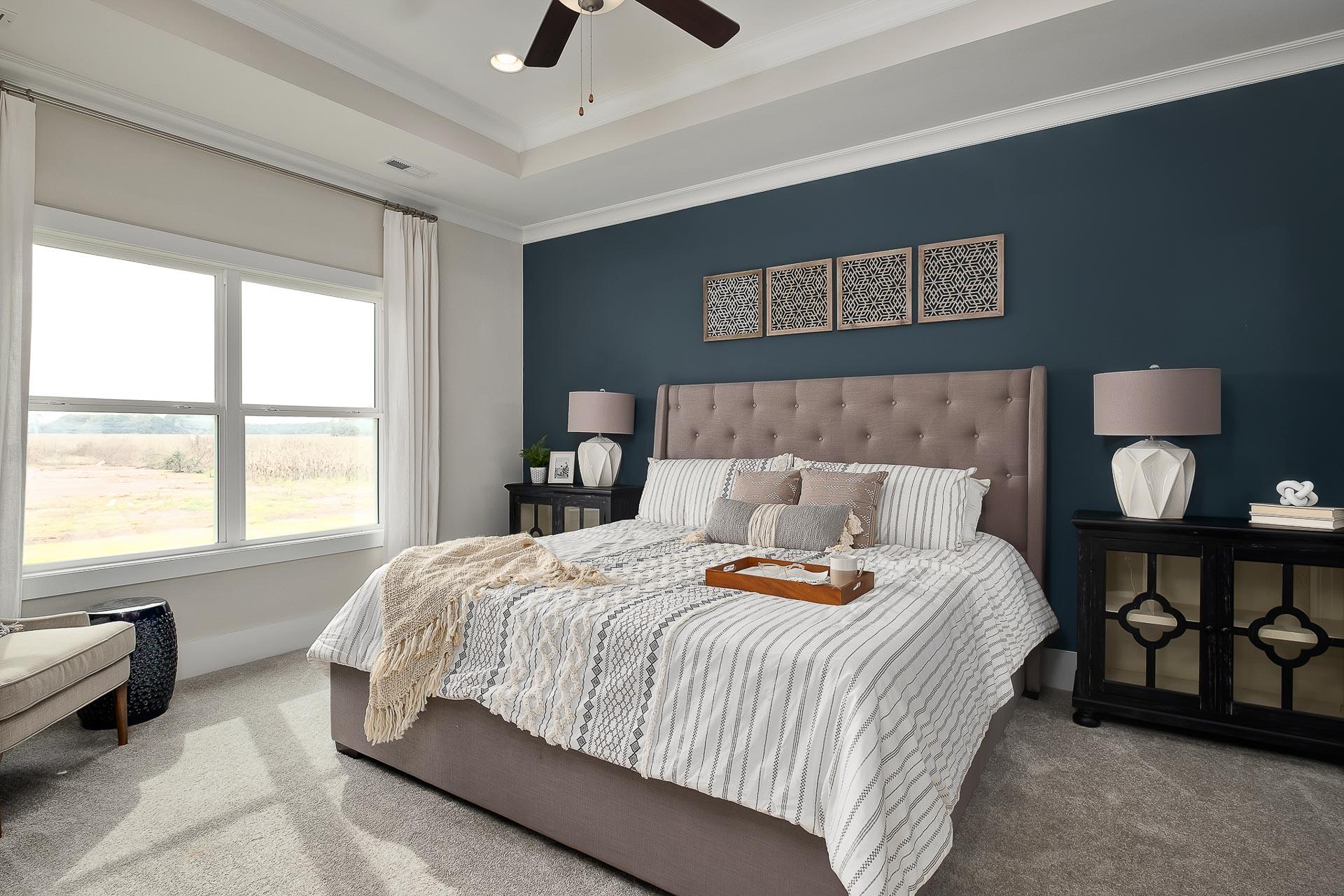 Spacious master bedroom in The Everett featuring navy accent wall, beige upholstered bed with striped duvet, ceiling fan, and large window