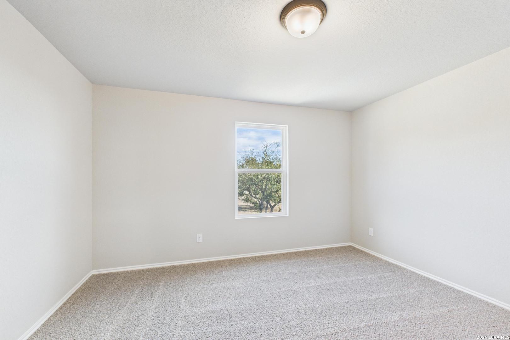 Bright empty bedroom with beige walls, carpeted floor, and large window overlooking trees in Davidson Homes The Douglas E, Bricewood, San Antonio