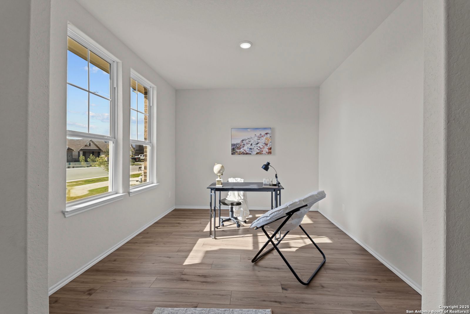 Bright home office with wooden desk, lamp, and abstract art on white walls, natural light via large windows in Davidson Homes The Douglas C, Seguin, Texas
