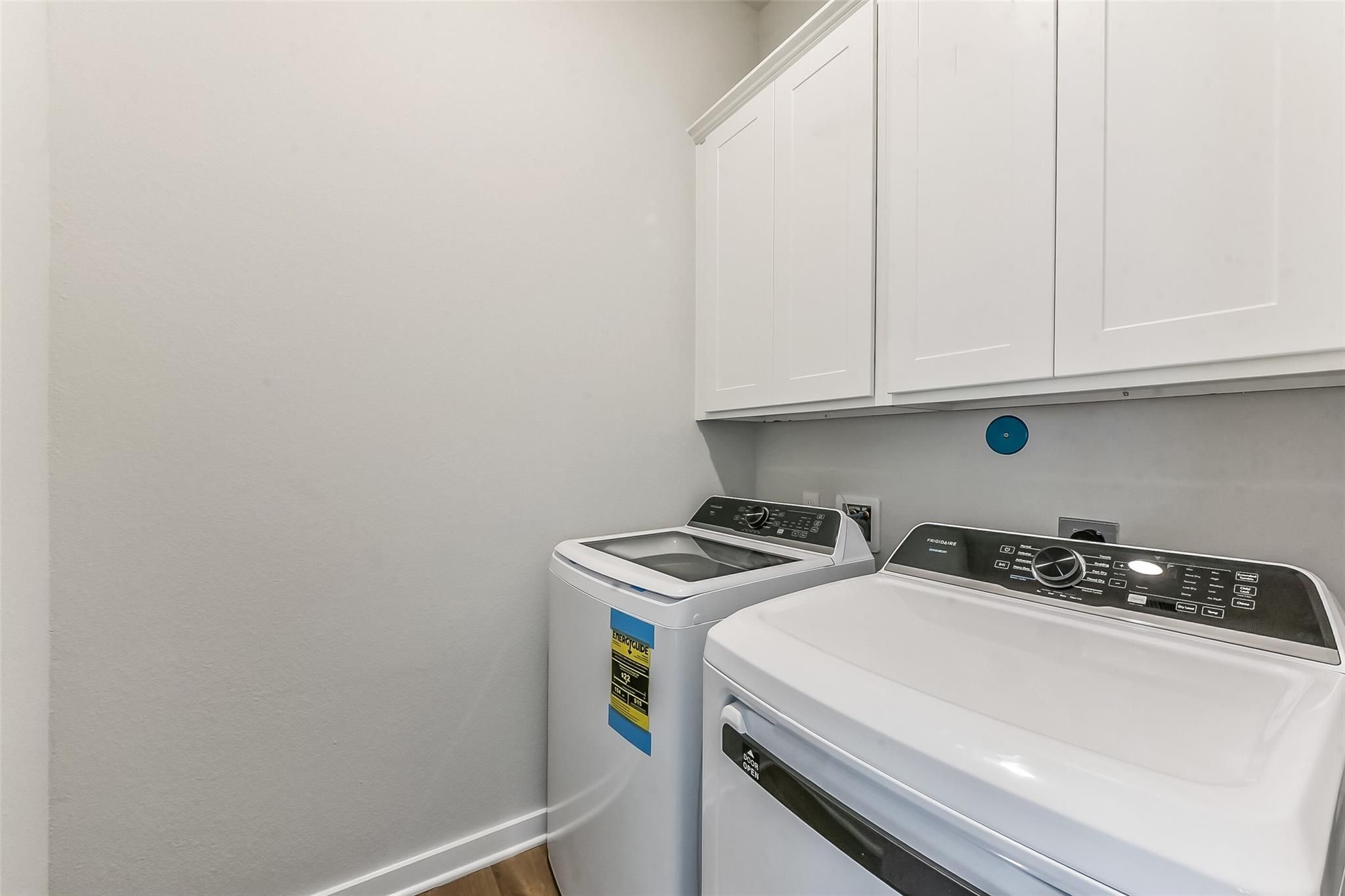 Modern laundry room featuring white front-load washer and dryer with overhead cabinets in Davidson Homes The Costa B, Dayton, Texas