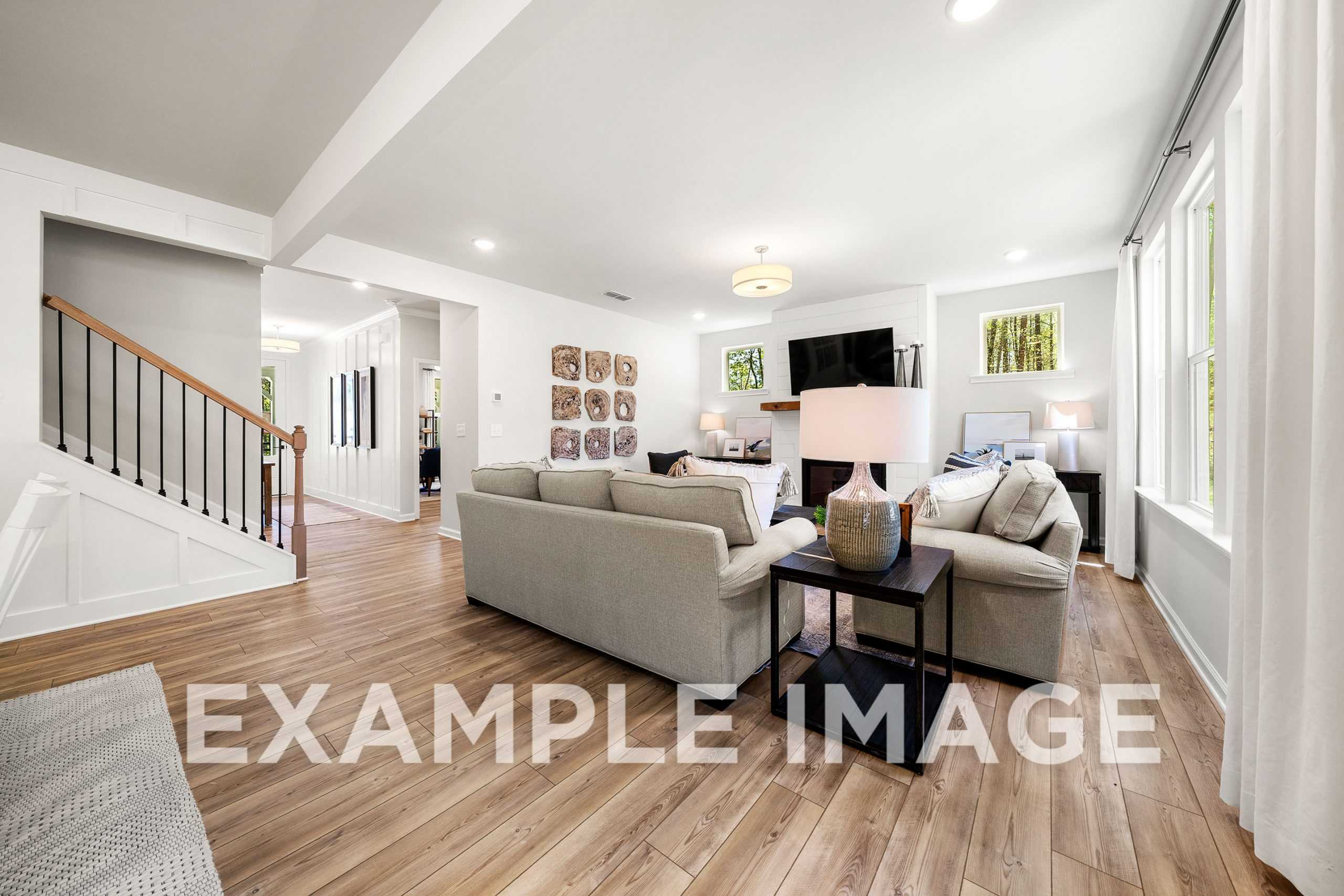 Spacious open-concept living room in The Hickory C with beige L-shaped sofa, mounted TV, wall art, lamps, hardwood floors, and staircase