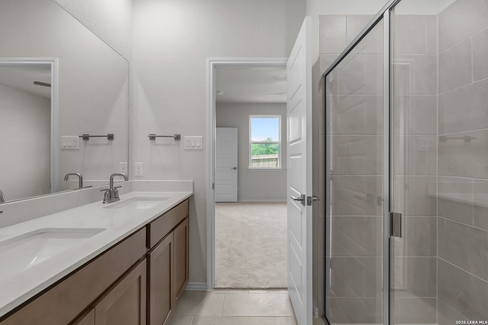 Modern bathroom with dual vanity, glass-enclosed shower, and bedroom view in The San Marcos F, Davidson Homes, San Antonio.